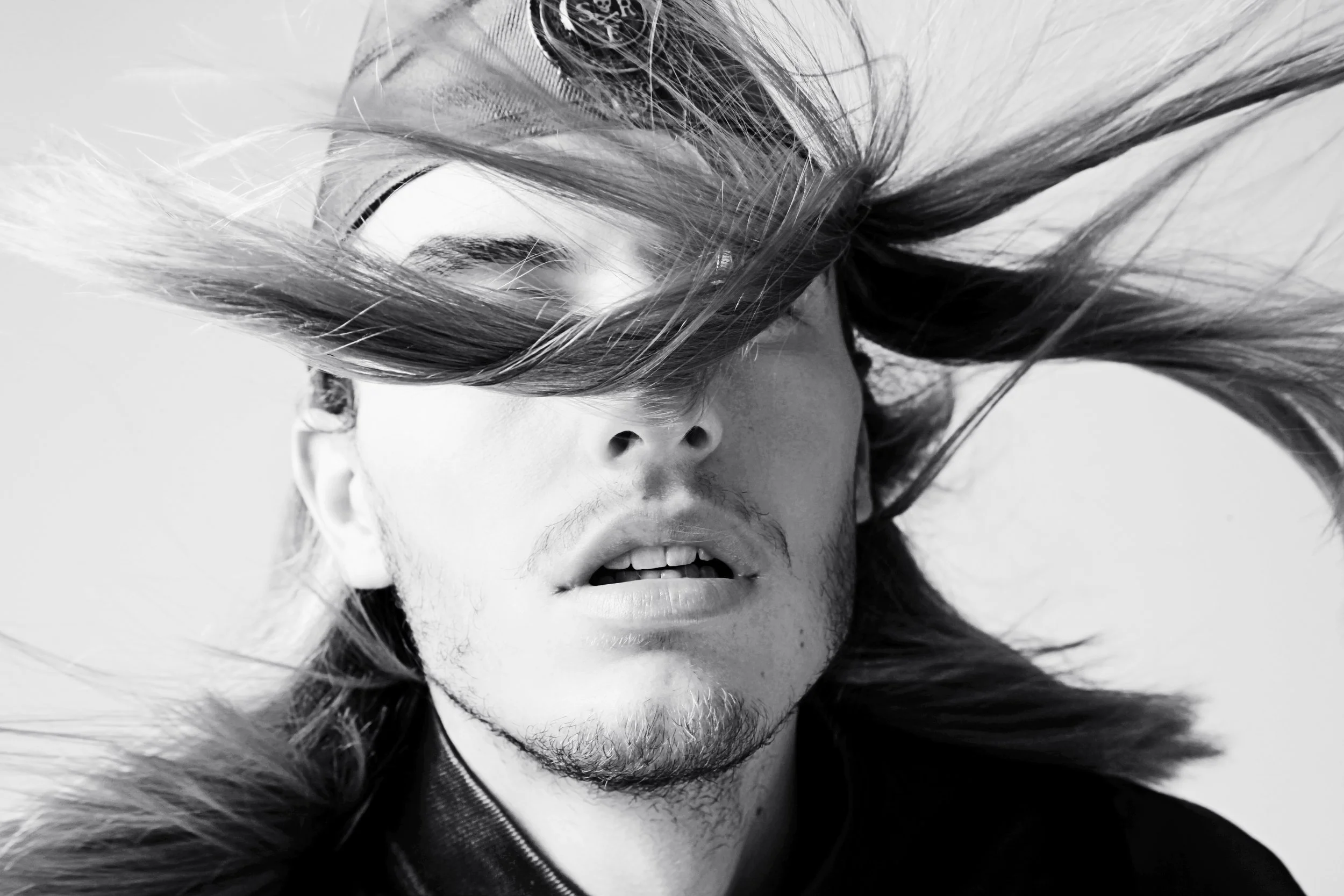 A black and white close-up photograph of a person's face with hair partially covering it. The person is wearing a fur-lined hood and has a piercing on their ear. Their lips are slightly parted, revealing some teeth.
