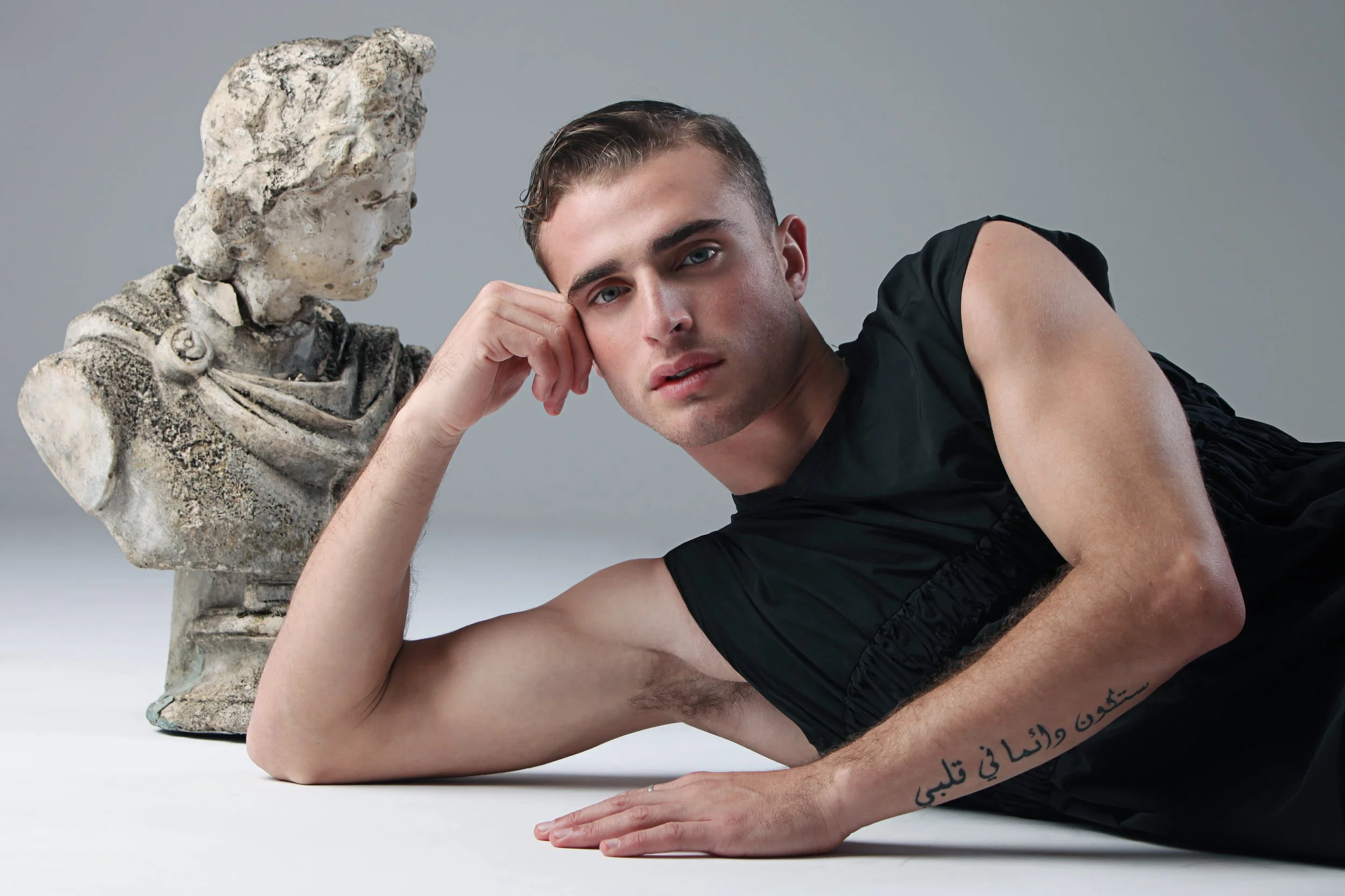 A young man with blue eyes and short brown hair reclining on the ground, resting his head on his left hand, next to a weathered classical marble bust of a woman. The man is wearing a black sleeveless shirt and has Arabic script tattooed on his right 