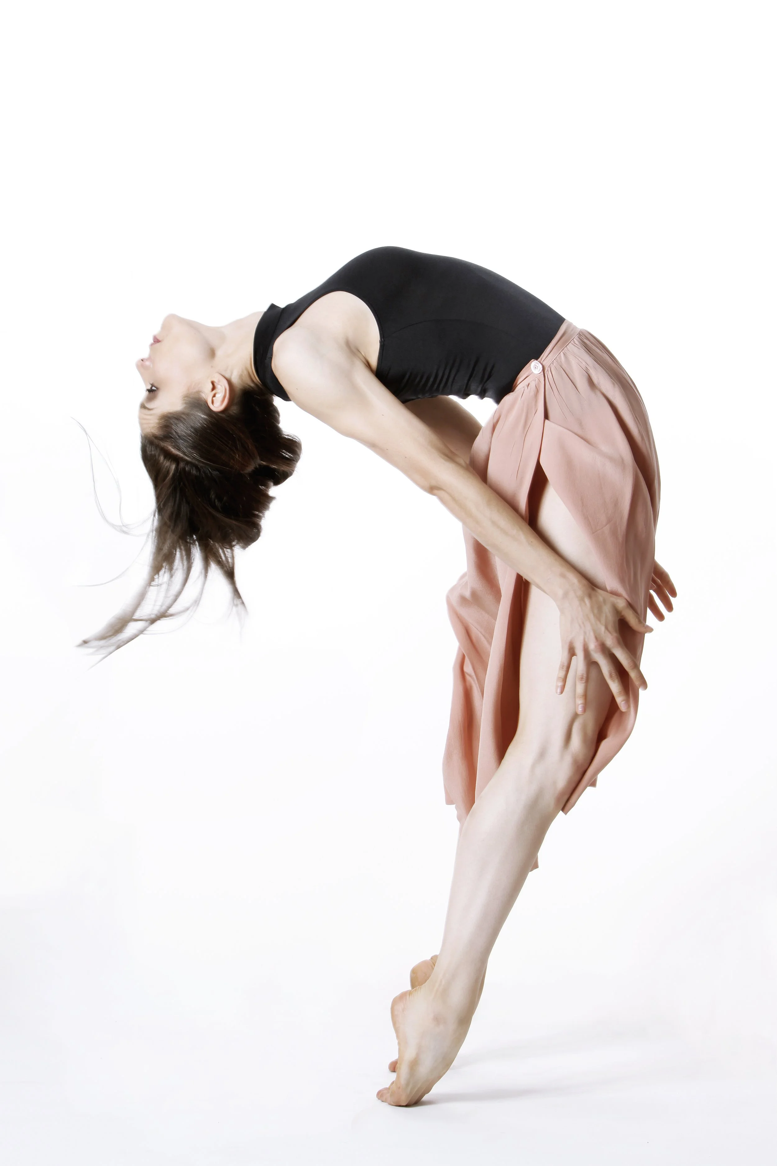 A woman wearing a black top and pink skirt performing a backbend pose with her head and hands on the ground and her right leg lifted, against a plain white background.