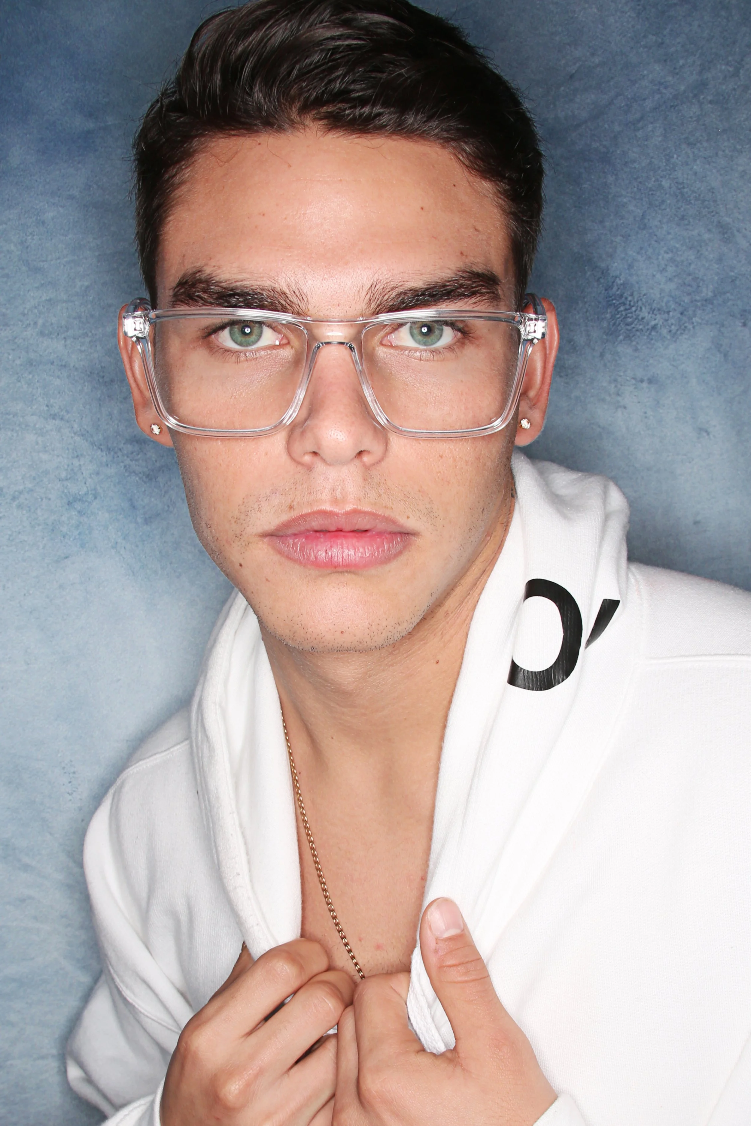 Close-up portrait of a young man with light skin, wearing clear glasses, a white hoodie, and a gold chain, partially unzipping the hoodie.