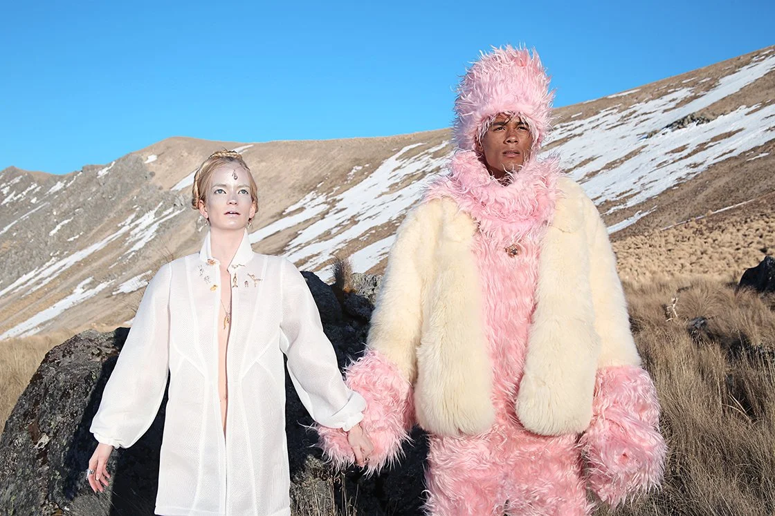 Two people standing outdoors in a mountainous landscape with patches of snow, holding hands, wearing elaborate fashion outfits. One person is dressed in a white, sheer dress with jewelry, and the other in a pink and cream furry costume with a large f
