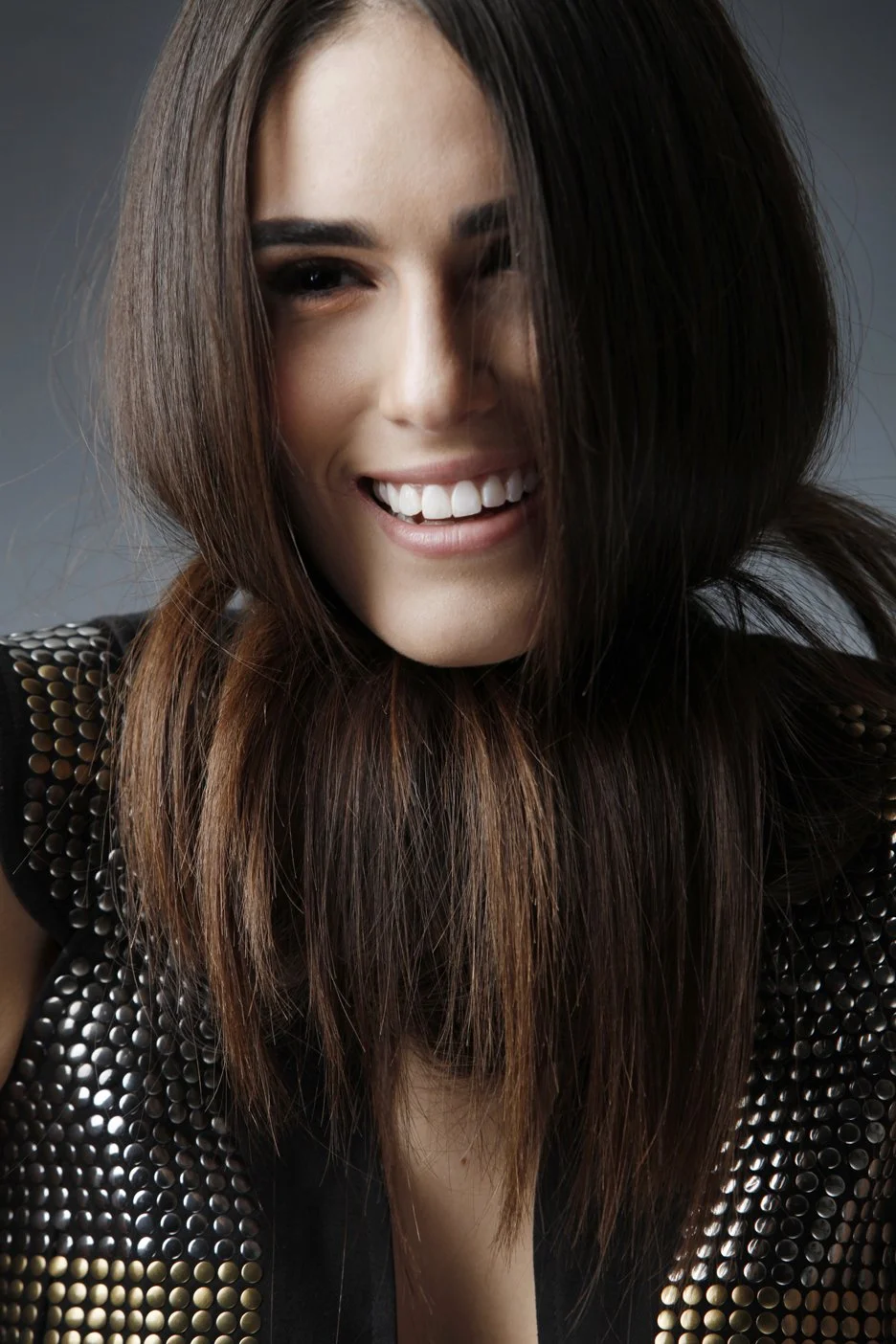 A young woman with long brown hair, smiling with teeth showing, wearing a black top with metallic embellishments.