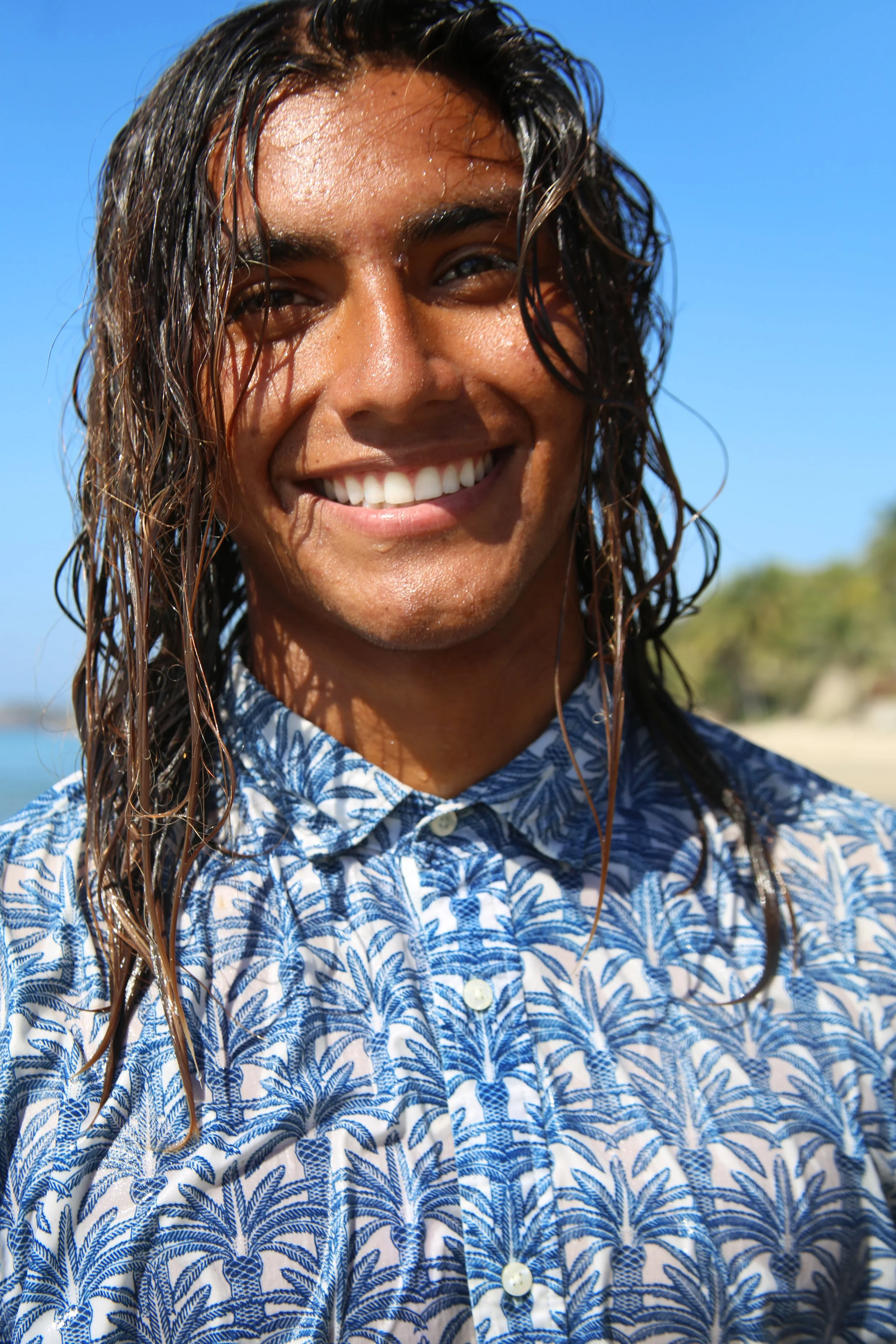 Close-up of a smiling man with wet, wavy hair, wearing a blue and white patterned shirt outdoors on a sunny day.