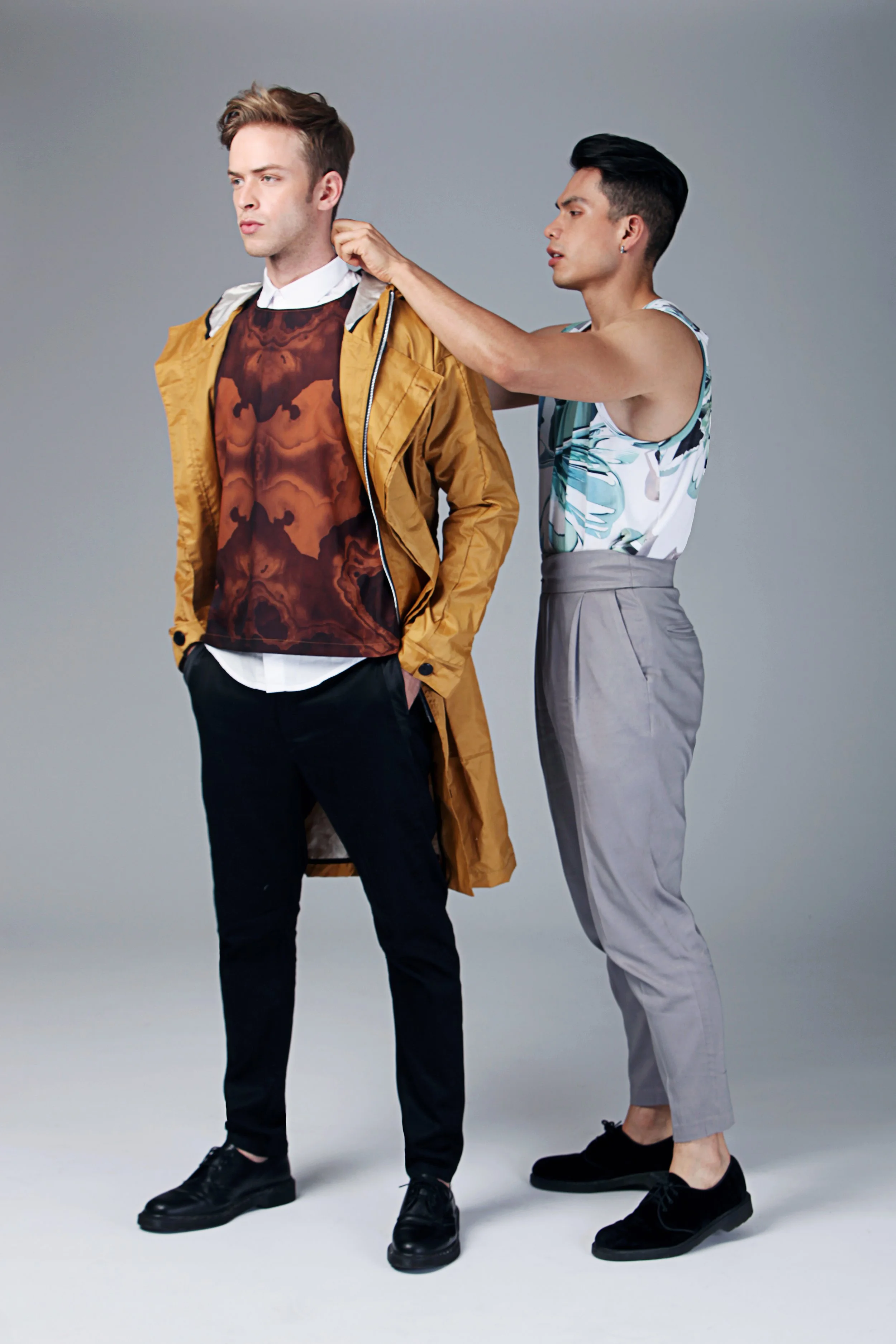 Two young men are standing against a plain gray background. The man on the left is wearing a mustard raincoat, black pants, and black shoes with a brown patterned shirt underneath. The man on the right is dressed in a sleeveless floral shirt, gray hi