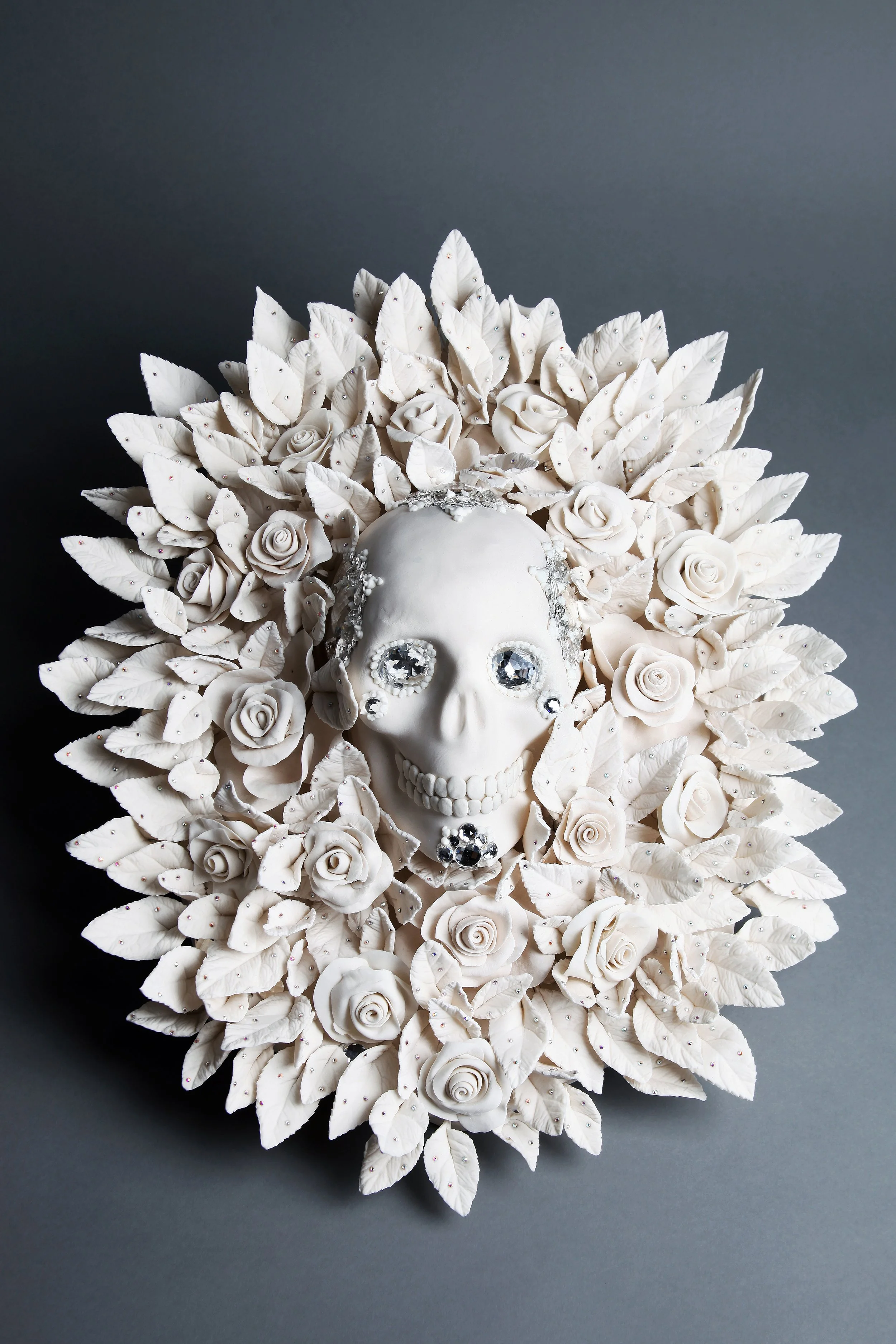 A decorative sculpture of a human skull adorned with an elaborate headdress made of white roses, leaves, and rhinestones, set against a gray background.