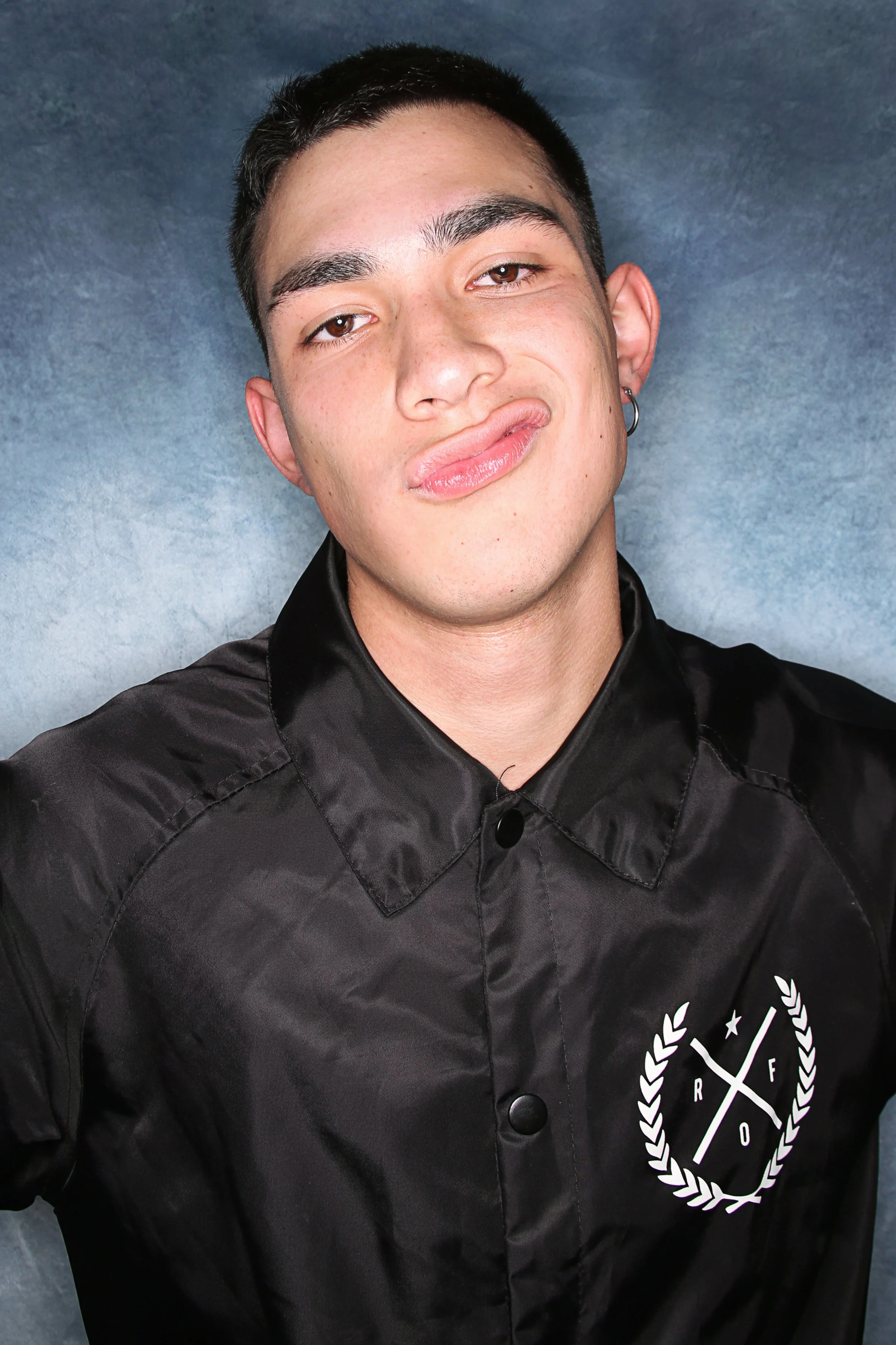 Close-up of a young man with short dark hair, light skin, and a mischievous expression, wearing a black button-up shirt with a white logo on the chest, standing against a gray textured background.