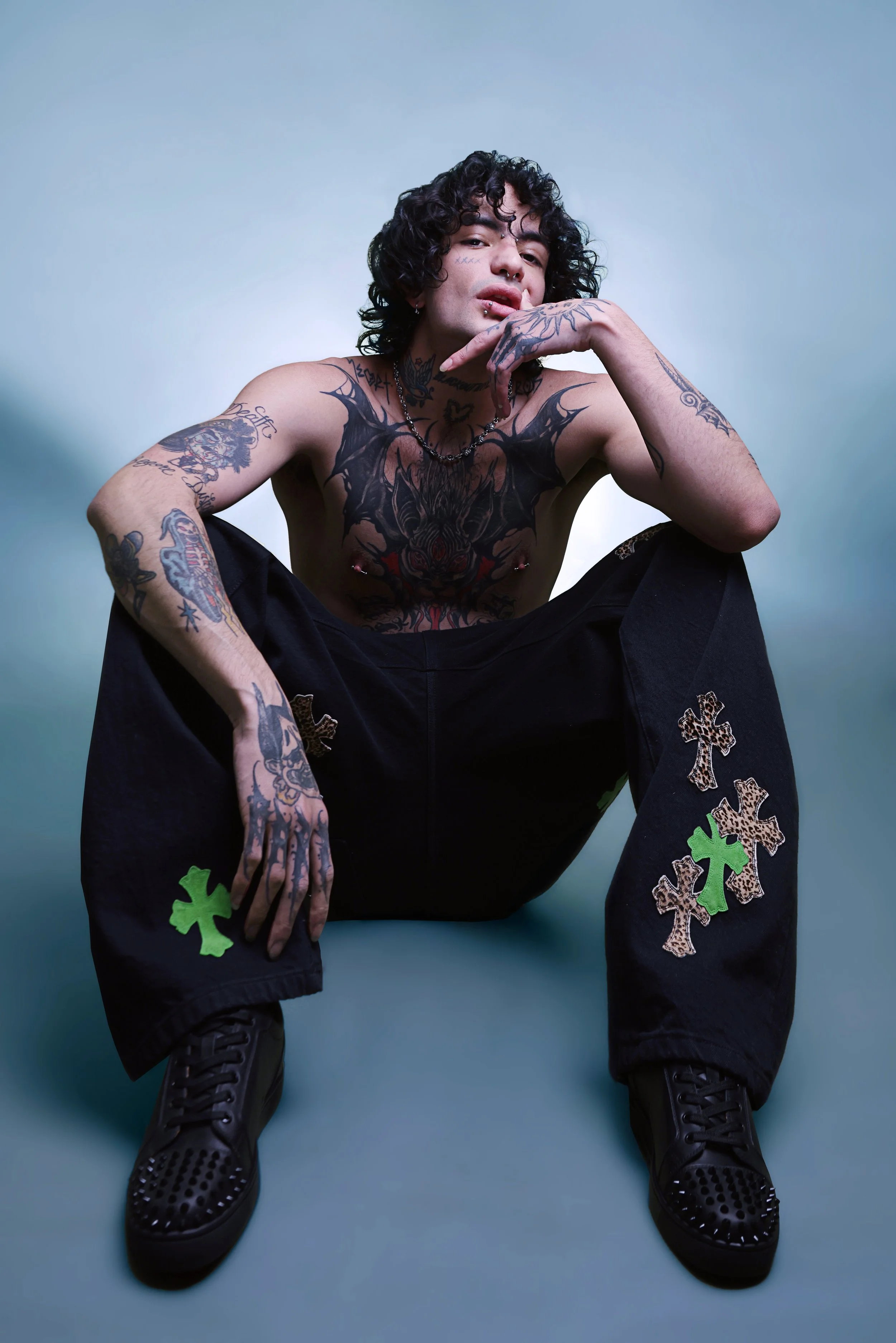 A young man with curly dark hair, tattoos on his upper body and arms, and piercings on his face. He is sitting with one knee up and his hand near his mouth, wearing black pants with green and leopard-print cross patches and black sneakers, against a 