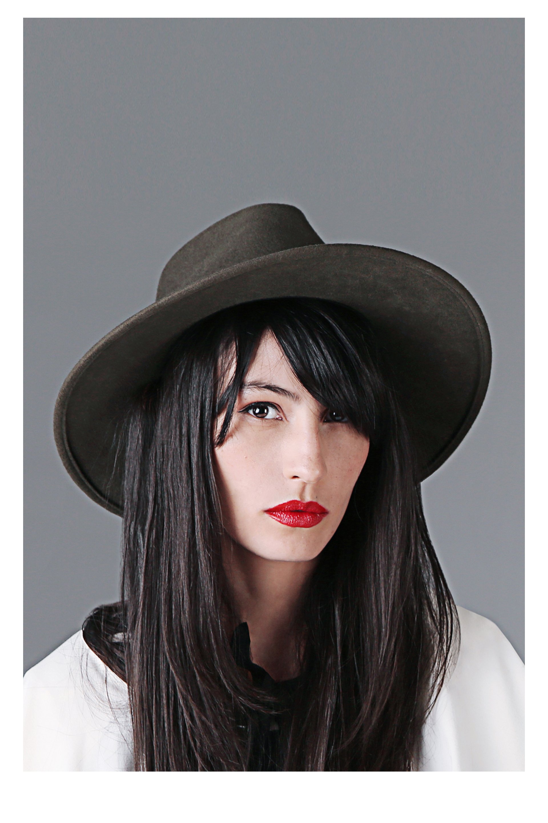 A woman with long black hair, wearing red lipstick and a wide-brimmed brown hat, and a white blazer over a black top, standing against a plain gray background.