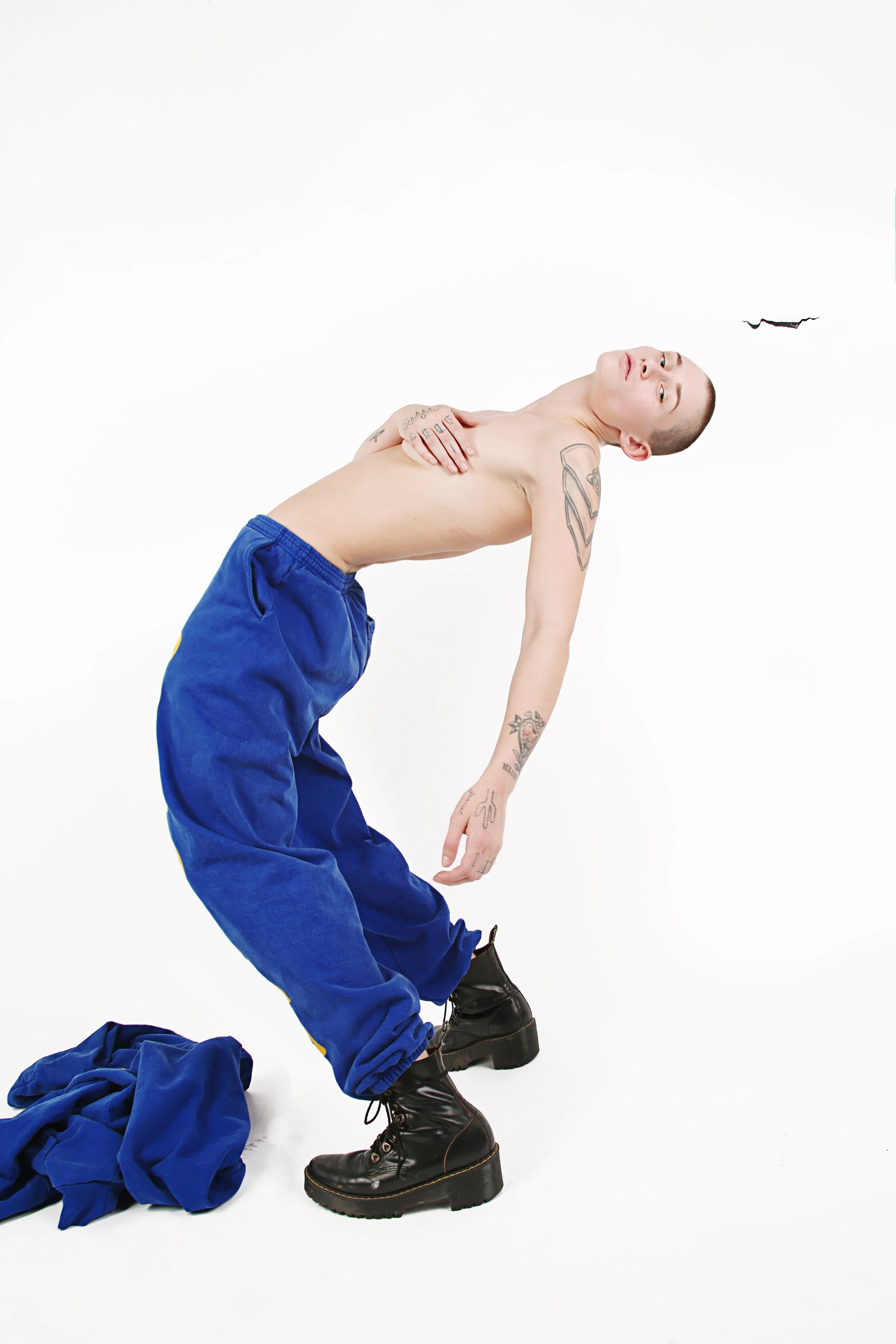 A young person with shaved head and tattoos posed against a white background, wearing black chunky boots, loose blue pants, and a shirtless upper body. Clothing is on the ground, and a small bat is flying nearby.