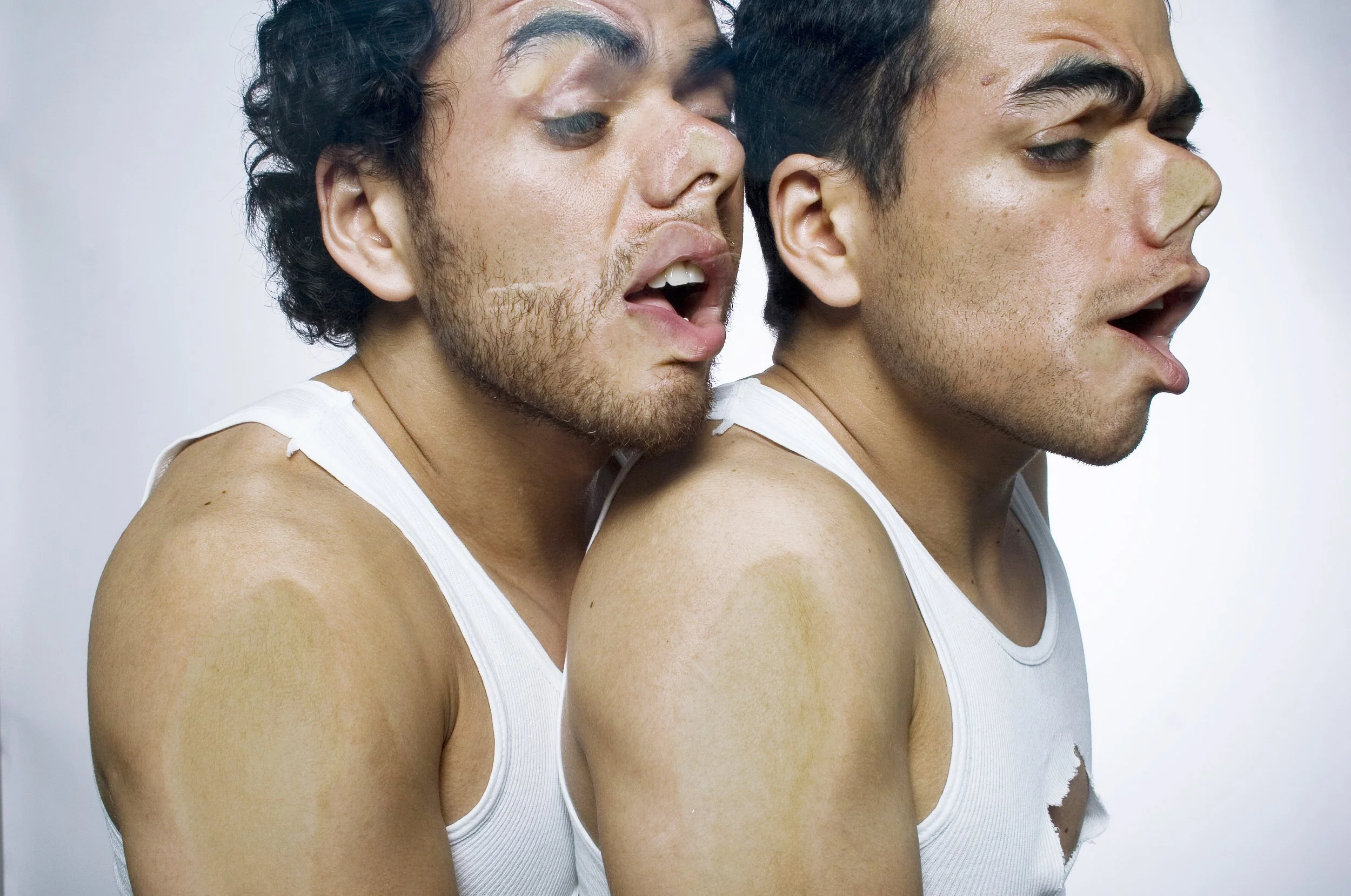 Two young men are standing side by side, wearing white tank tops, with one having curly black hair and the other having straight dark hair. The man on the left is slightly behind the man on the right, and both have their eyes closed or looking down. 