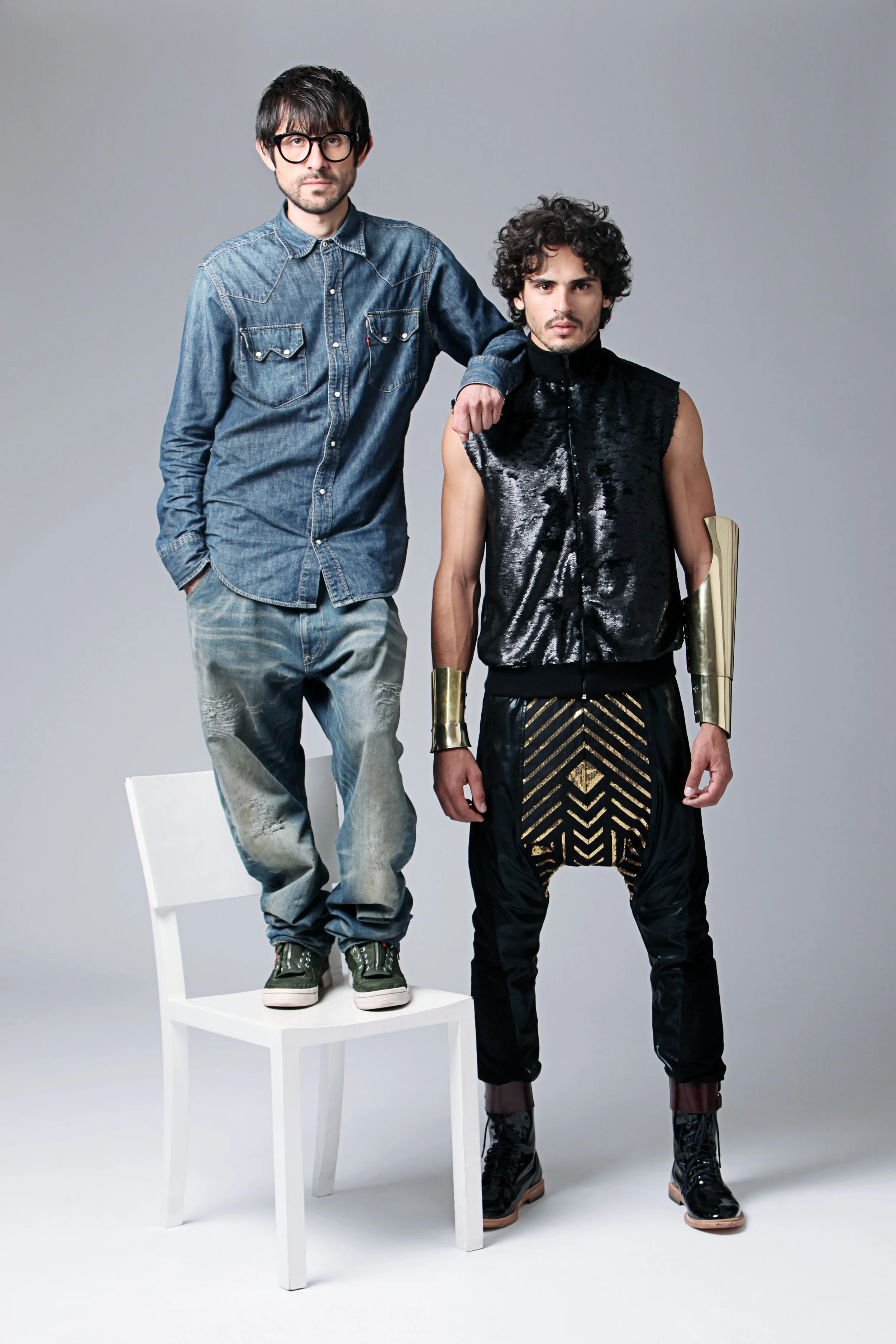 Two men in fashionable clothing pose in a studio with a neutral background. One man stands on a white chair with one hand in his pocket, wearing a denim shirt and jeans. The other man stands beside him, dressed in a shiny, black, sleeveless jacket an