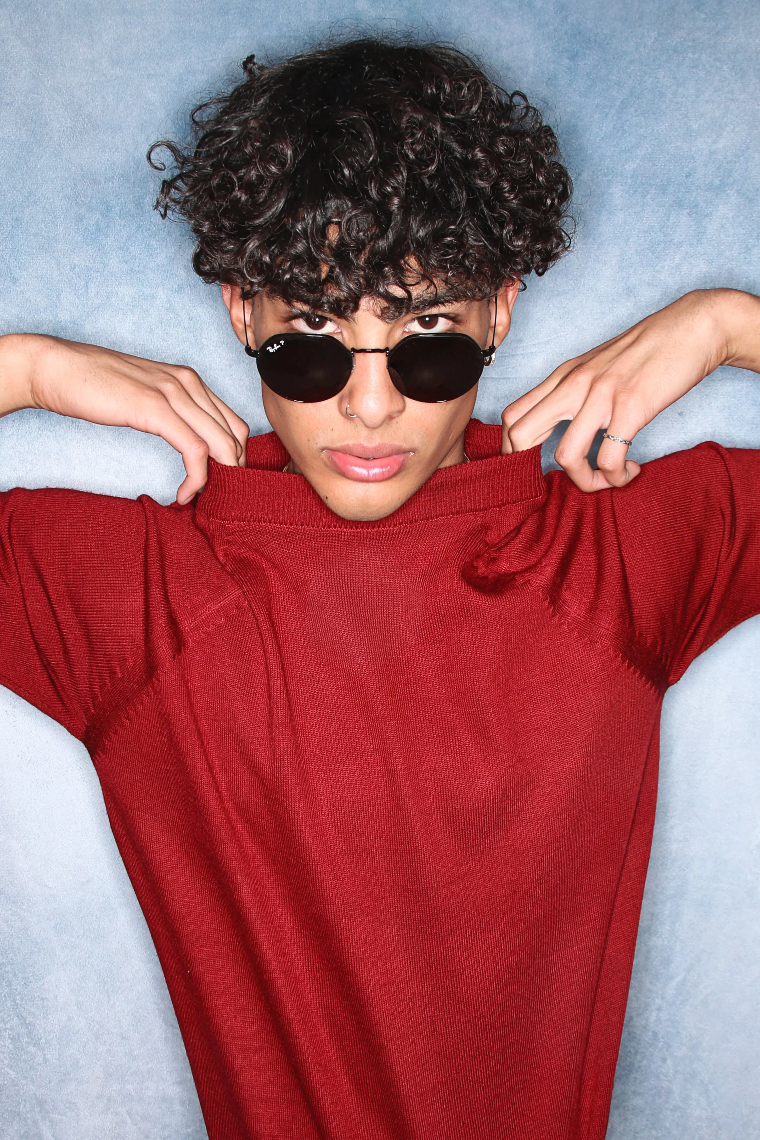 Young person with curly dark hair wearing black sunglasses and a red hoodie, lying against a blue surface.
