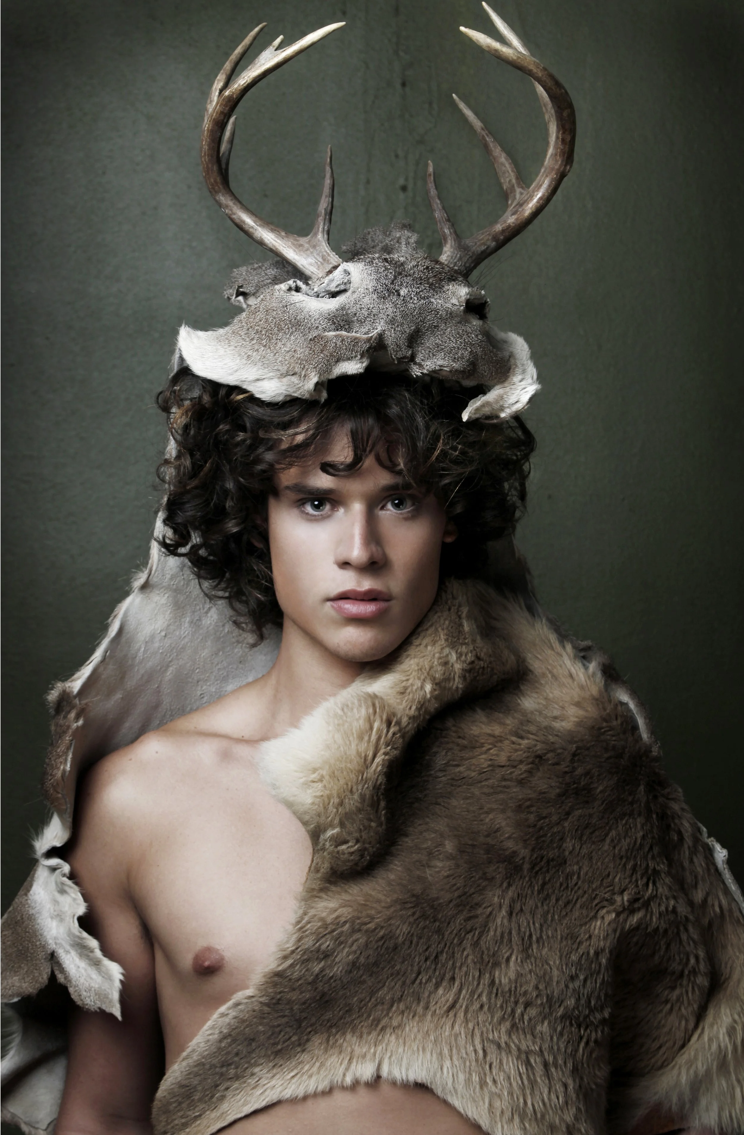 A person with curly hair and blue eyes, with a taxidermy deer head wearing antlers on top of their head, and a fur coat draped over their shoulders, against a dark green background.