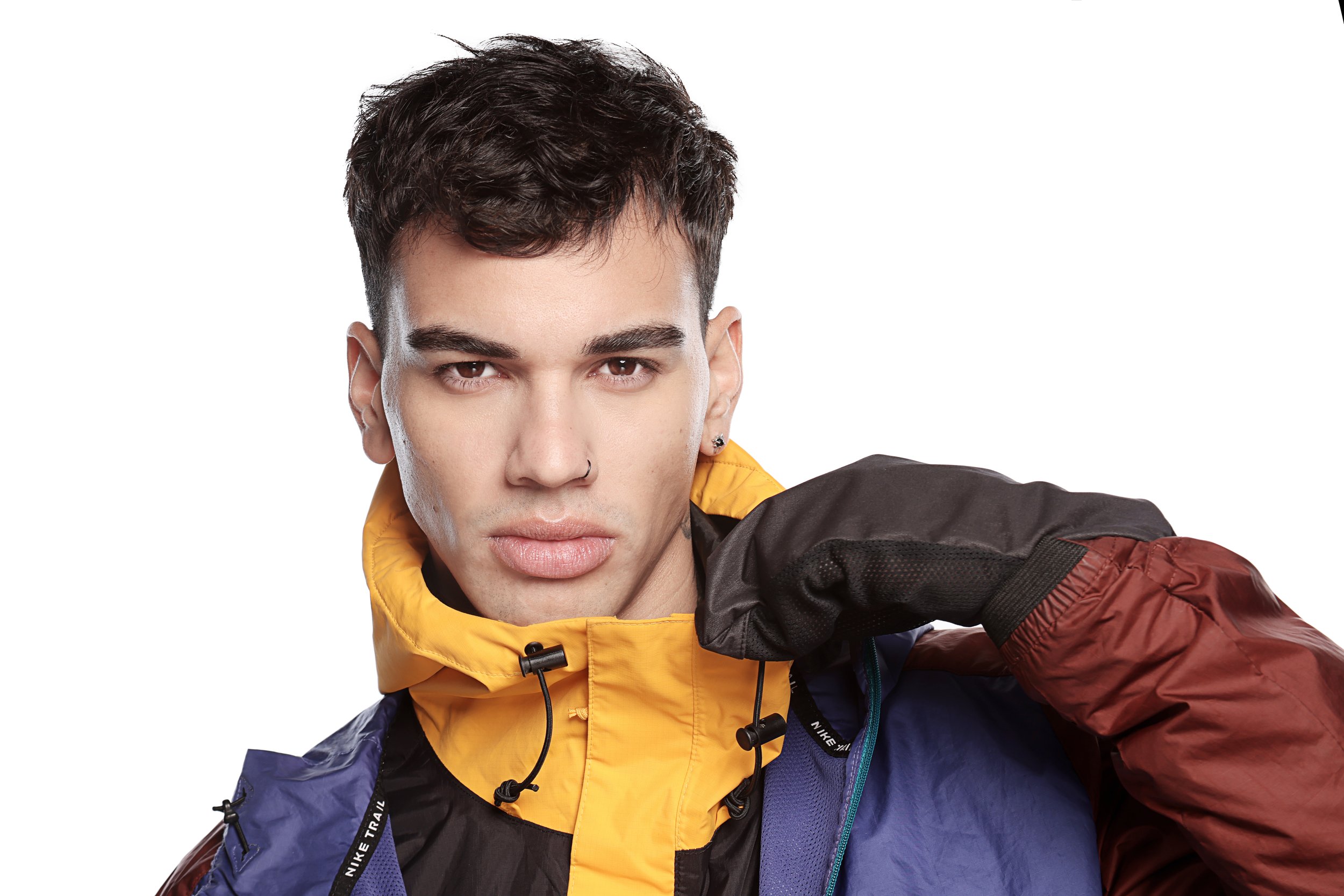 Young man with styled dark hair, wearing a multicolored jacket with a yellow collar, and looking directly at the camera against a plain white background.