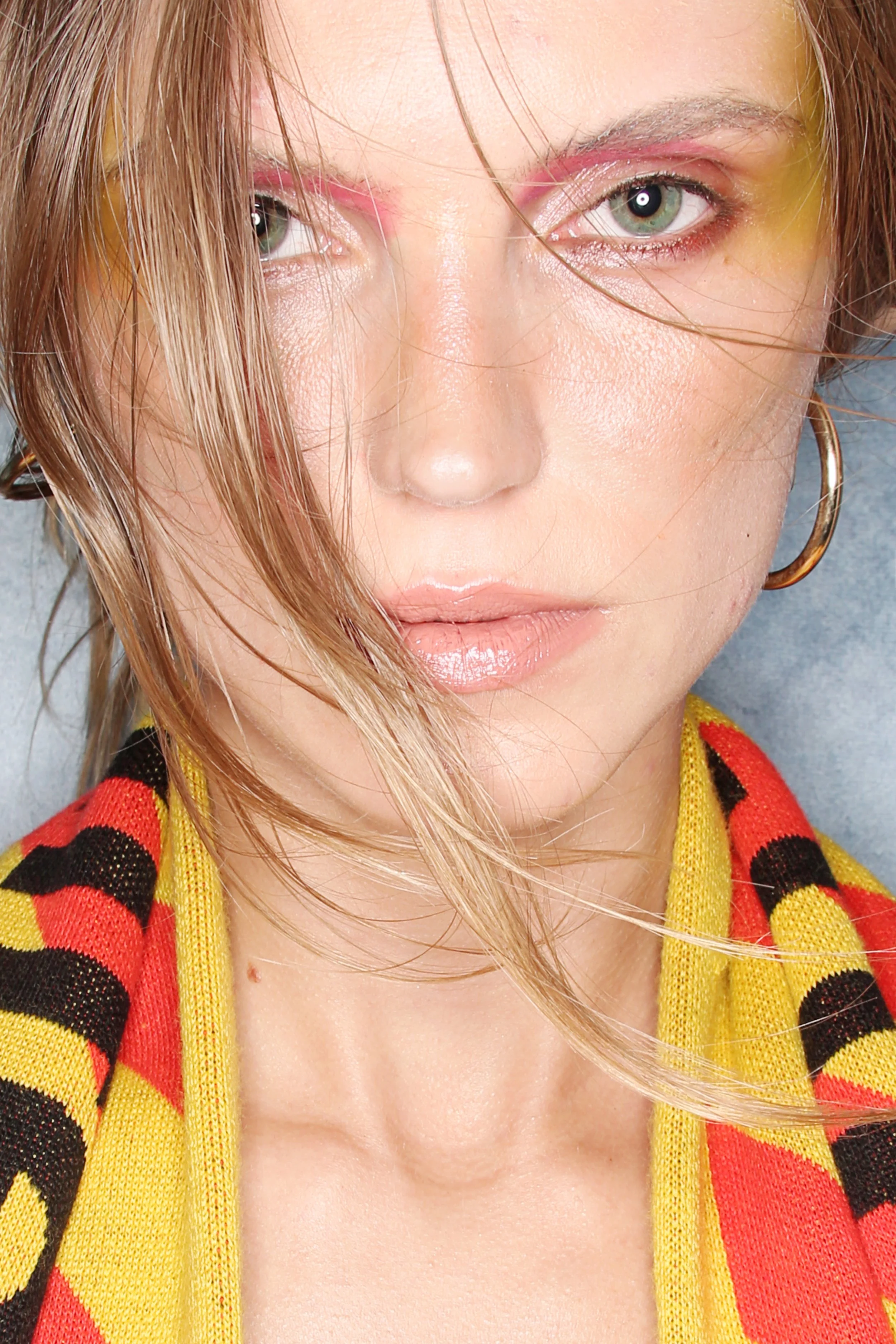 Close-up of a woman with green eyes, light makeup, and light brown hair, wearing a red and yellow striped garment.