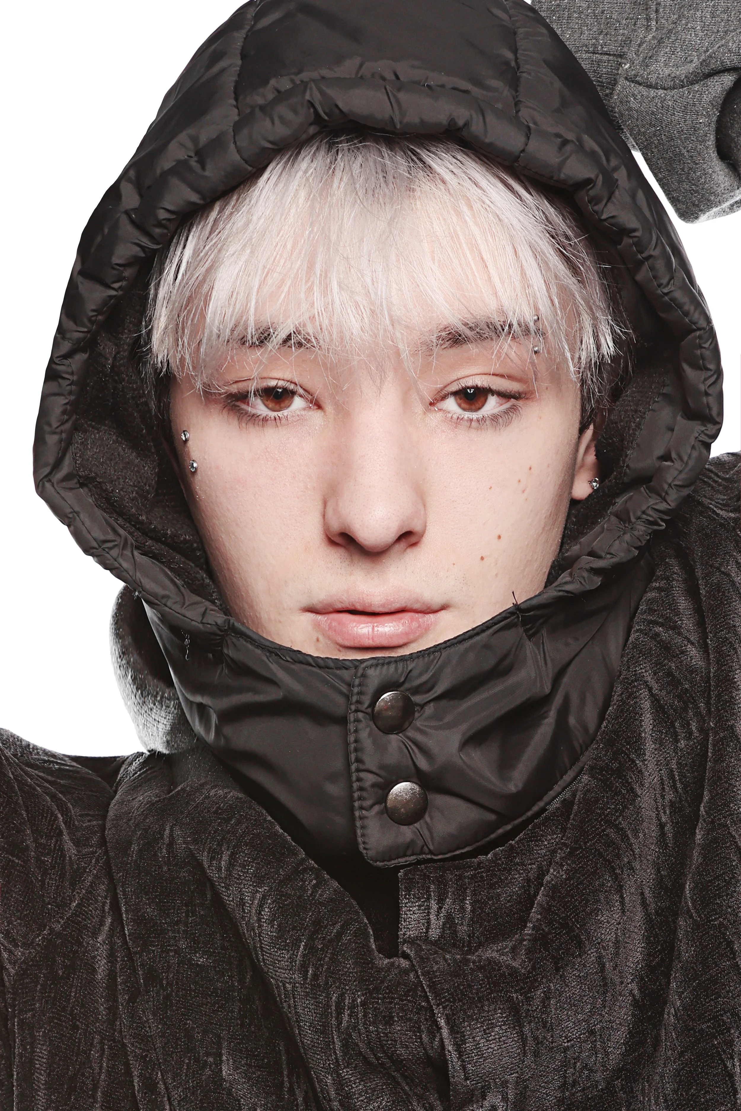 A close-up of a young person with platinum blonde hair, light skin, wearing a black hooded jacket with a shiny exterior, and a gray sweater underneath.