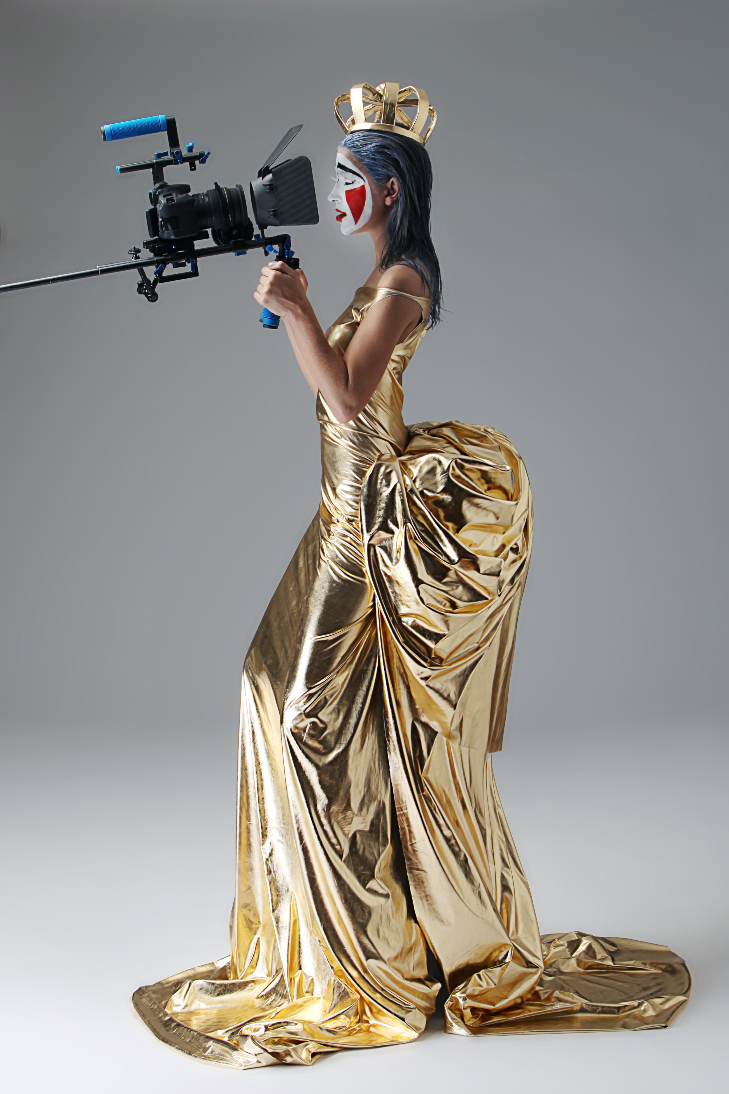 A woman dressed in a shiny gold gown with a dramatic train, wearing a gold crown and theatrical face makeup with bold red, black, and white patterns, is filming with a professional camera.