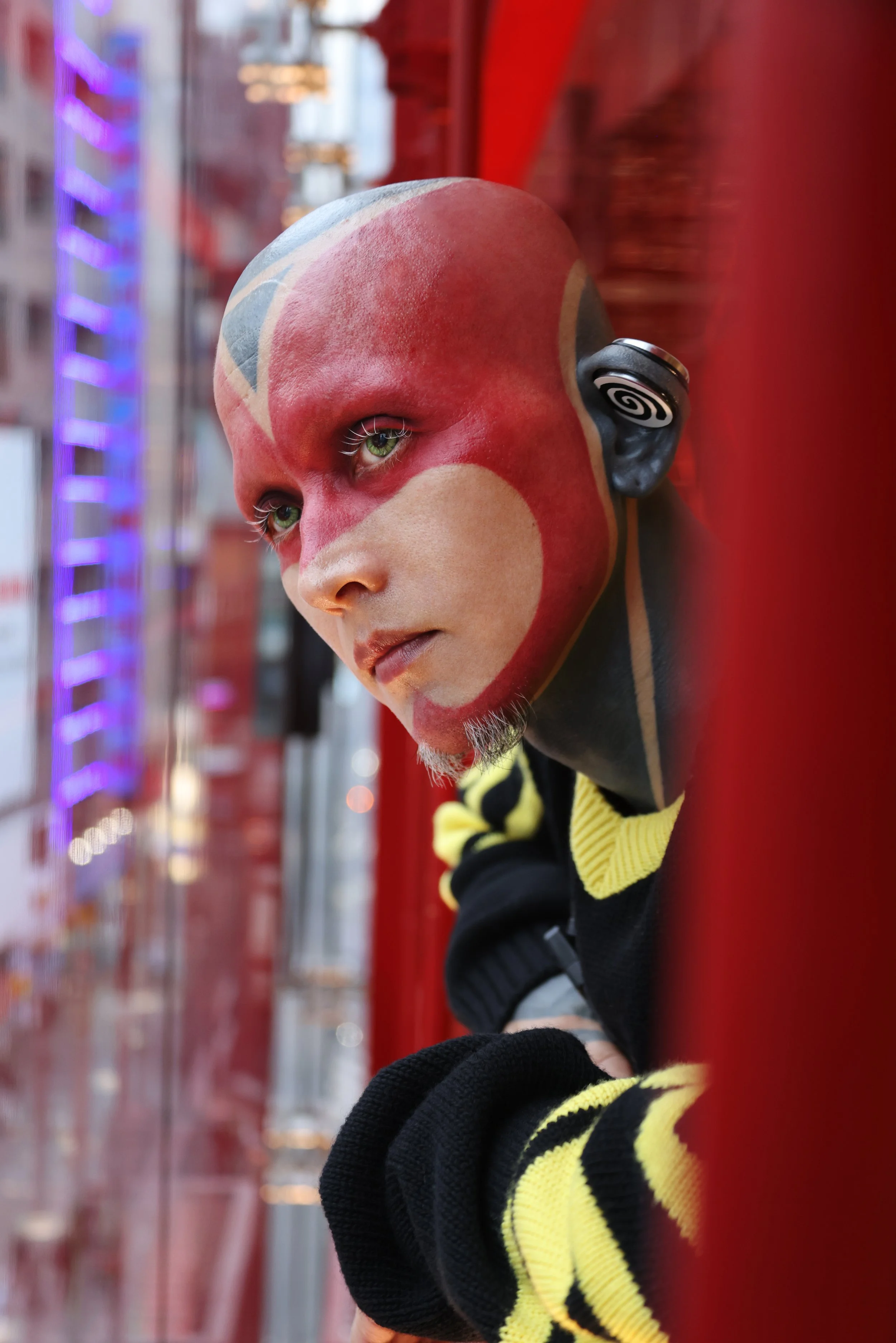 A person with face paint and green eyes looking sideways, wearing a black and yellow striped sweater, standing against a red background with buildings and neon lights in the background.