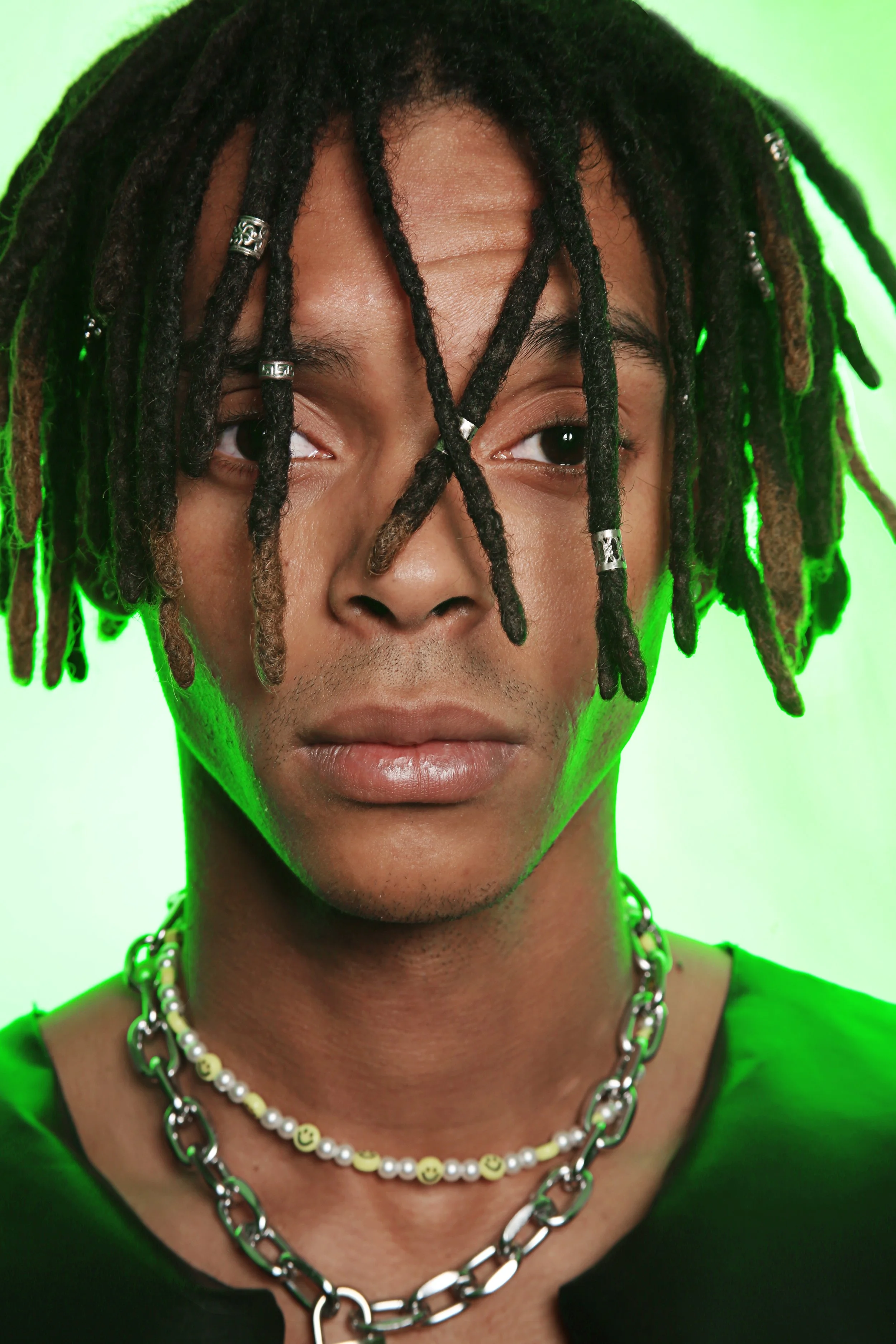 A close-up portrait of a man with dreadlocks, wearing layered silver and beaded necklaces, against a green background.