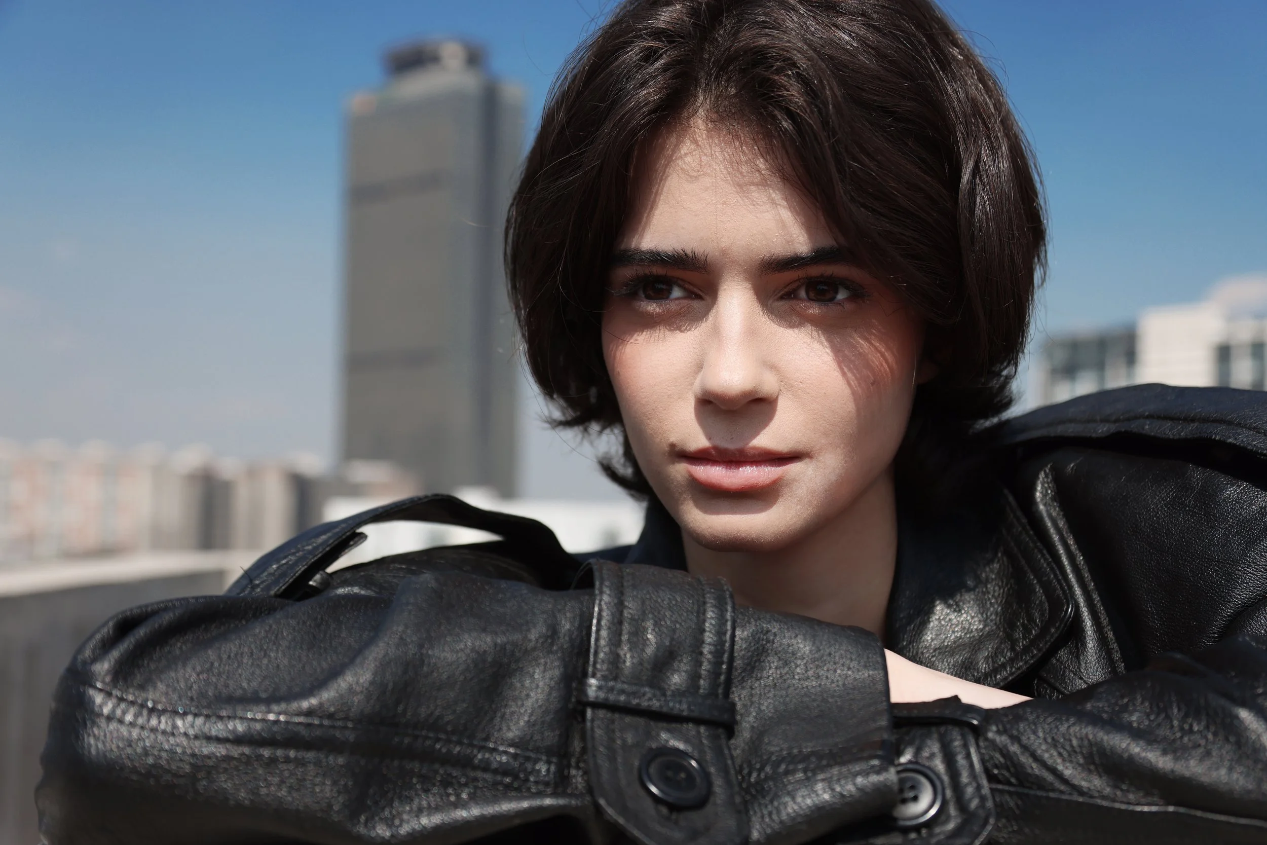 Close-up of a woman with short brown hair and brown eyes wearing a black leather jacket, leaning on her arm on a rooftop with a city skyline and a tall building in the background.
