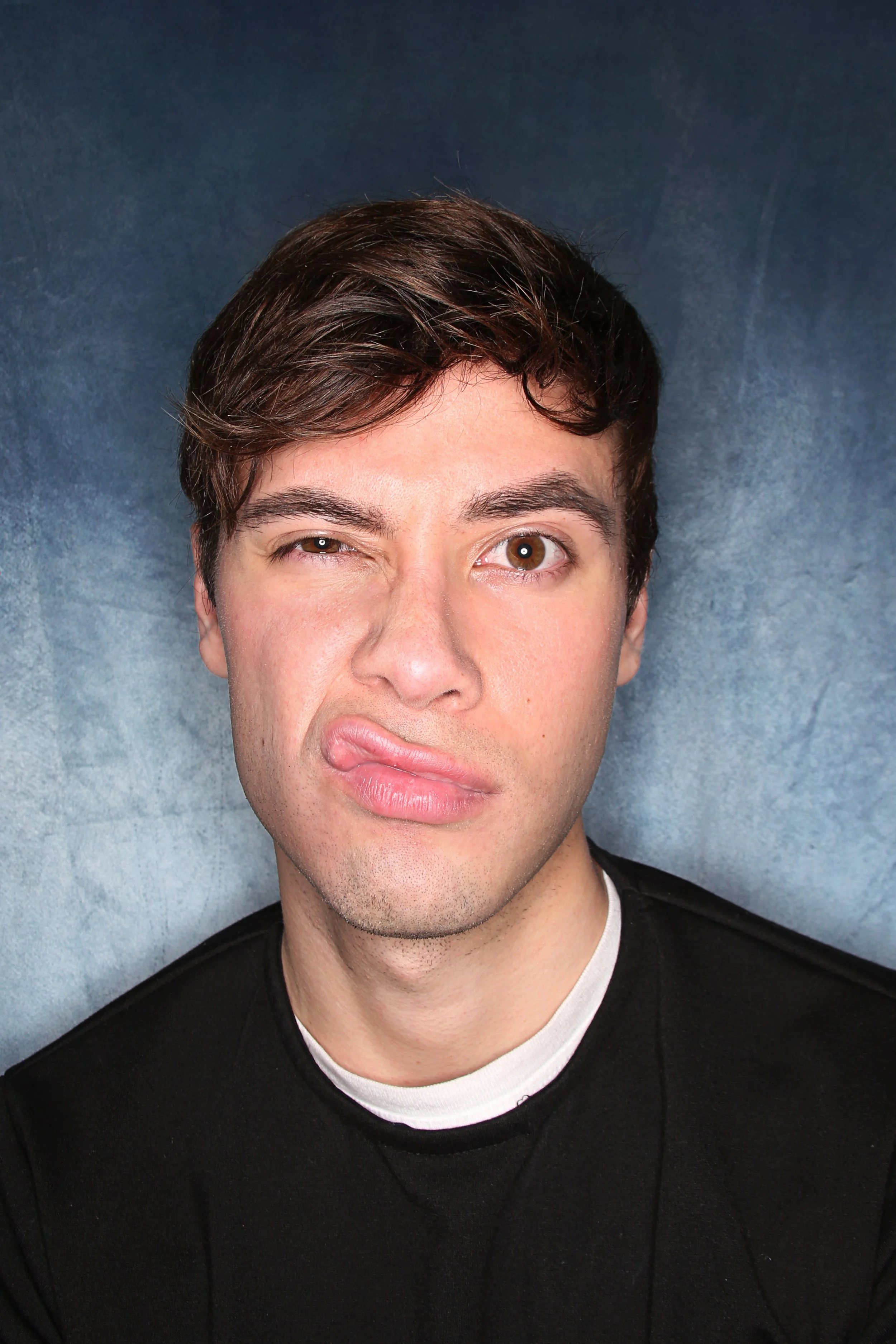A man with short brown hair winking and making a funny face, wearing a black shirt with a white undershirt, standing against a textured blue background.
