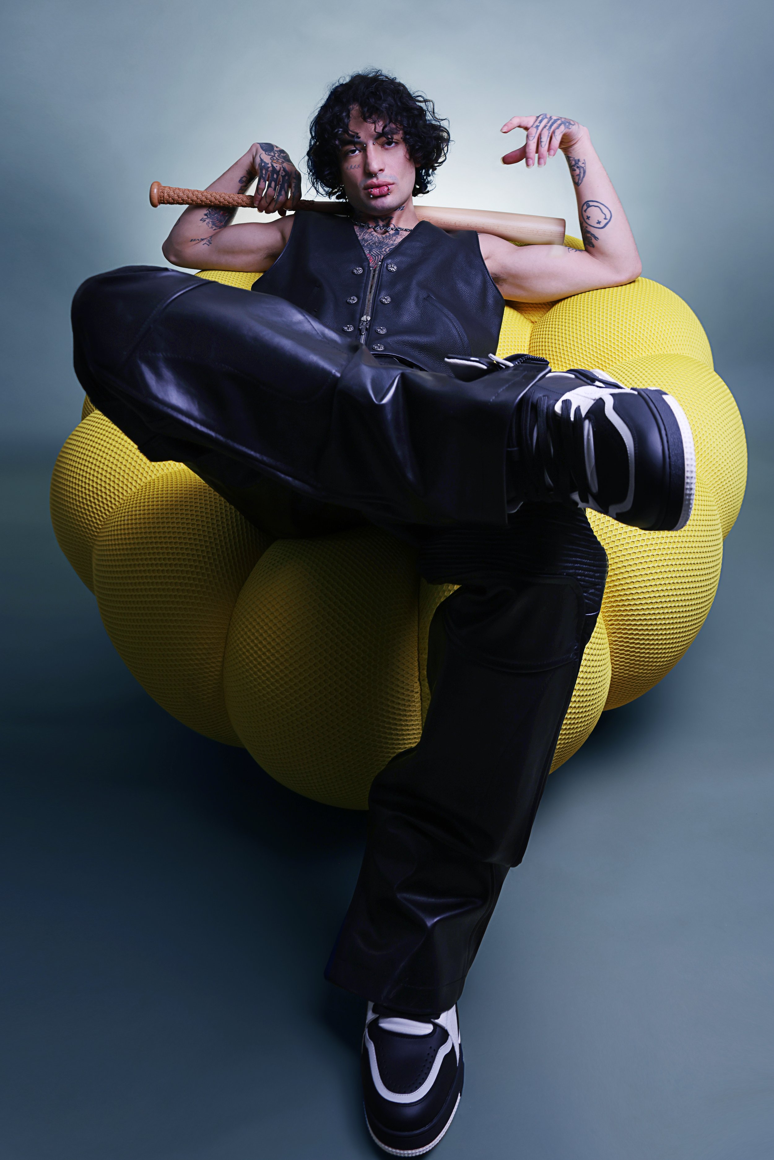Young man in black leather vest and pants sitting on a yellow modern armchair, holding a baseball bat resting on his shoulders, with tattoos, curly black hair, and sneakers, against a plain background.