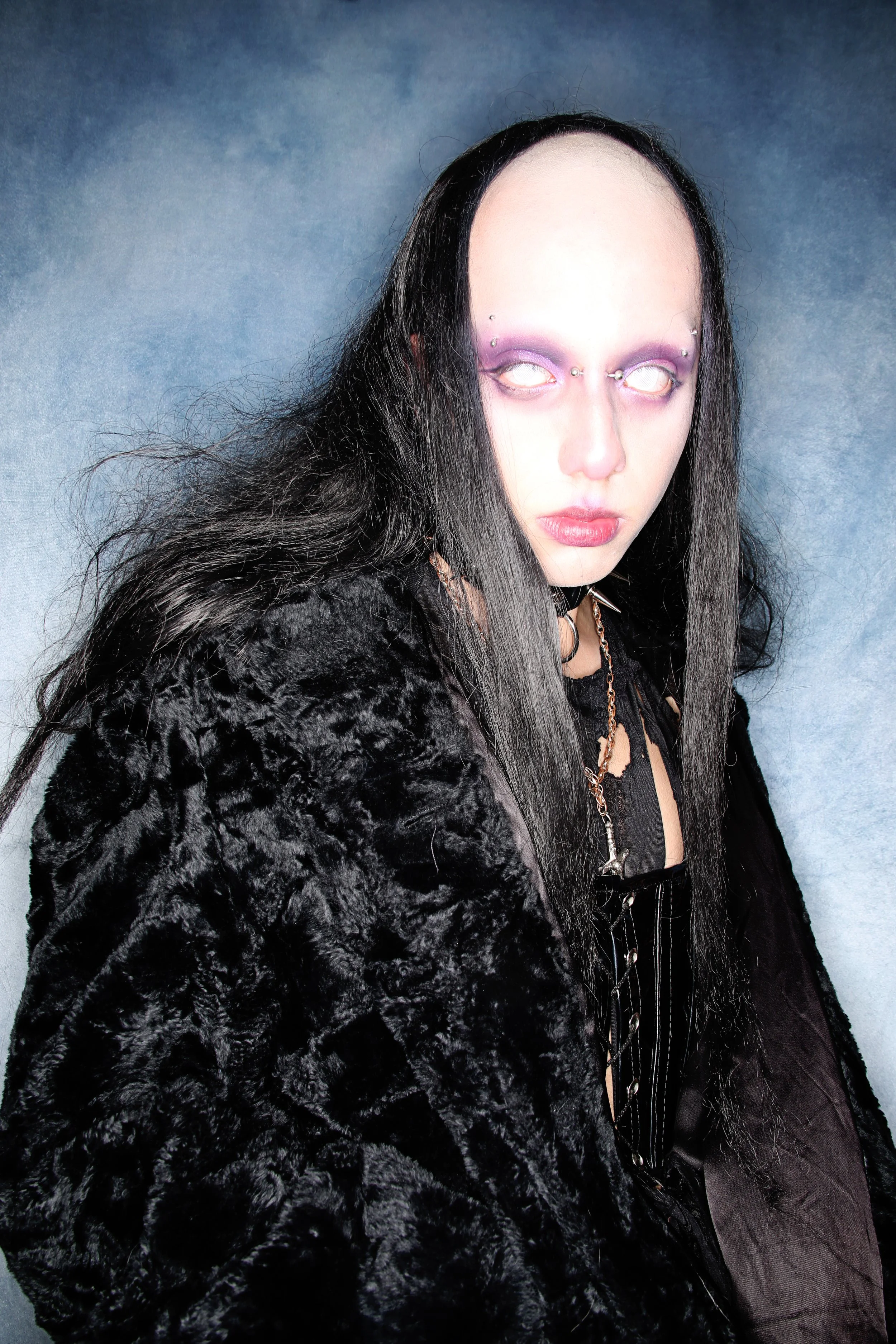 A person with long black hair and pale skin, wearing gothic style makeup with purple eyeshadow, pierced eyebrows, and white contact lenses. They are dressed in a black velvet fur coat and a black lace top, accessorized with chains and spikes, against