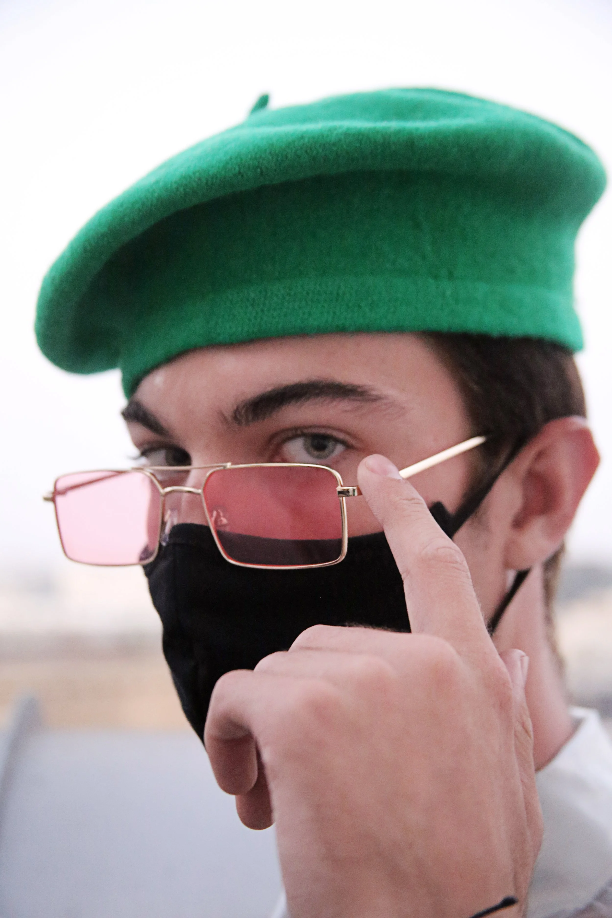 Close-up of a person's face wearing a green beret, pink-tinted sunglasses, and a black face mask, with their finger touching the side of the glasses.