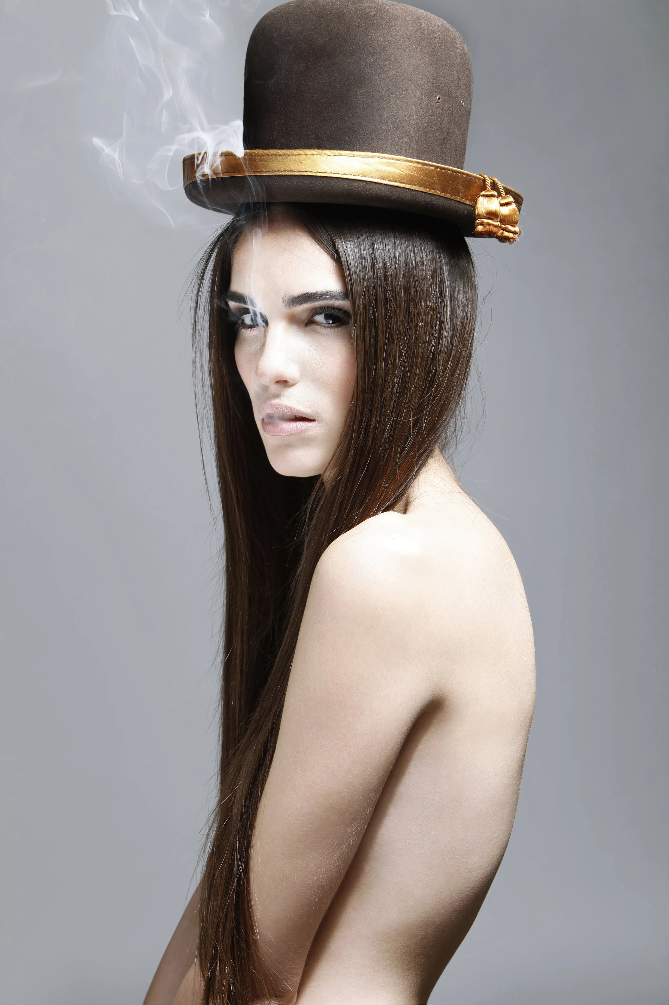 A woman with long brown hair and minimal makeup is seen from her shoulder up, wearing a large brown top hat with a gold band and a small tassel, against a plain gray background.