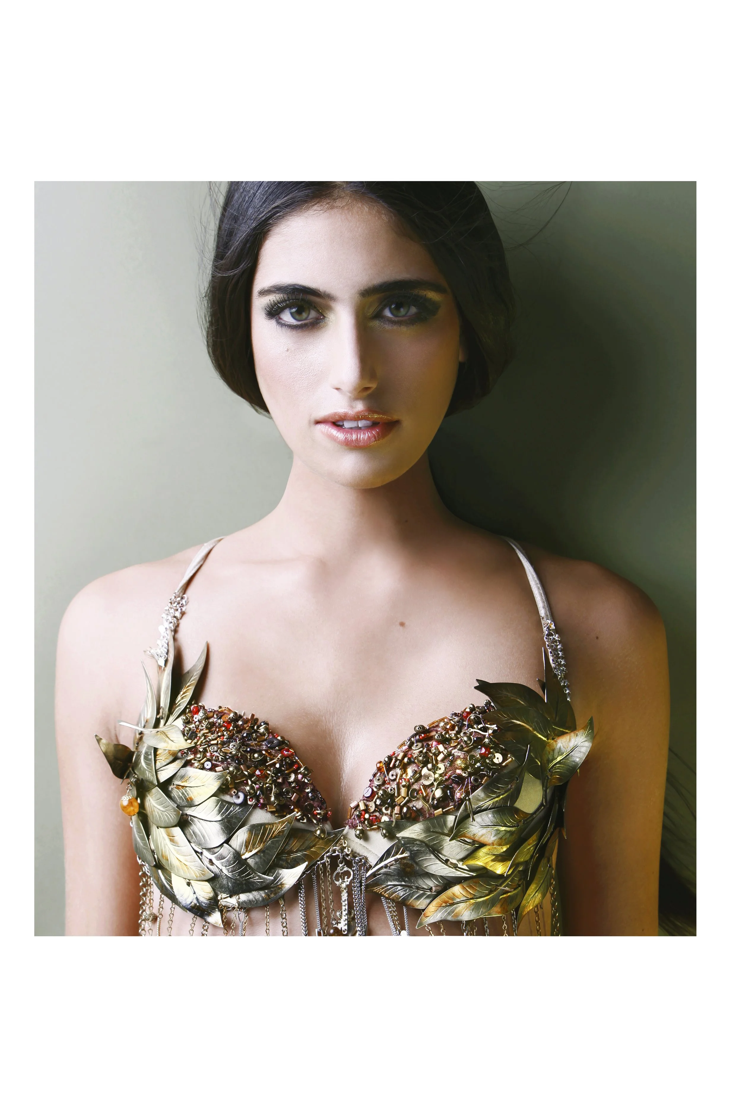 A young woman with short black hair and green eyes, wearing a dress decorated with gold leaves, beads, and chains, standing against a plain wall.