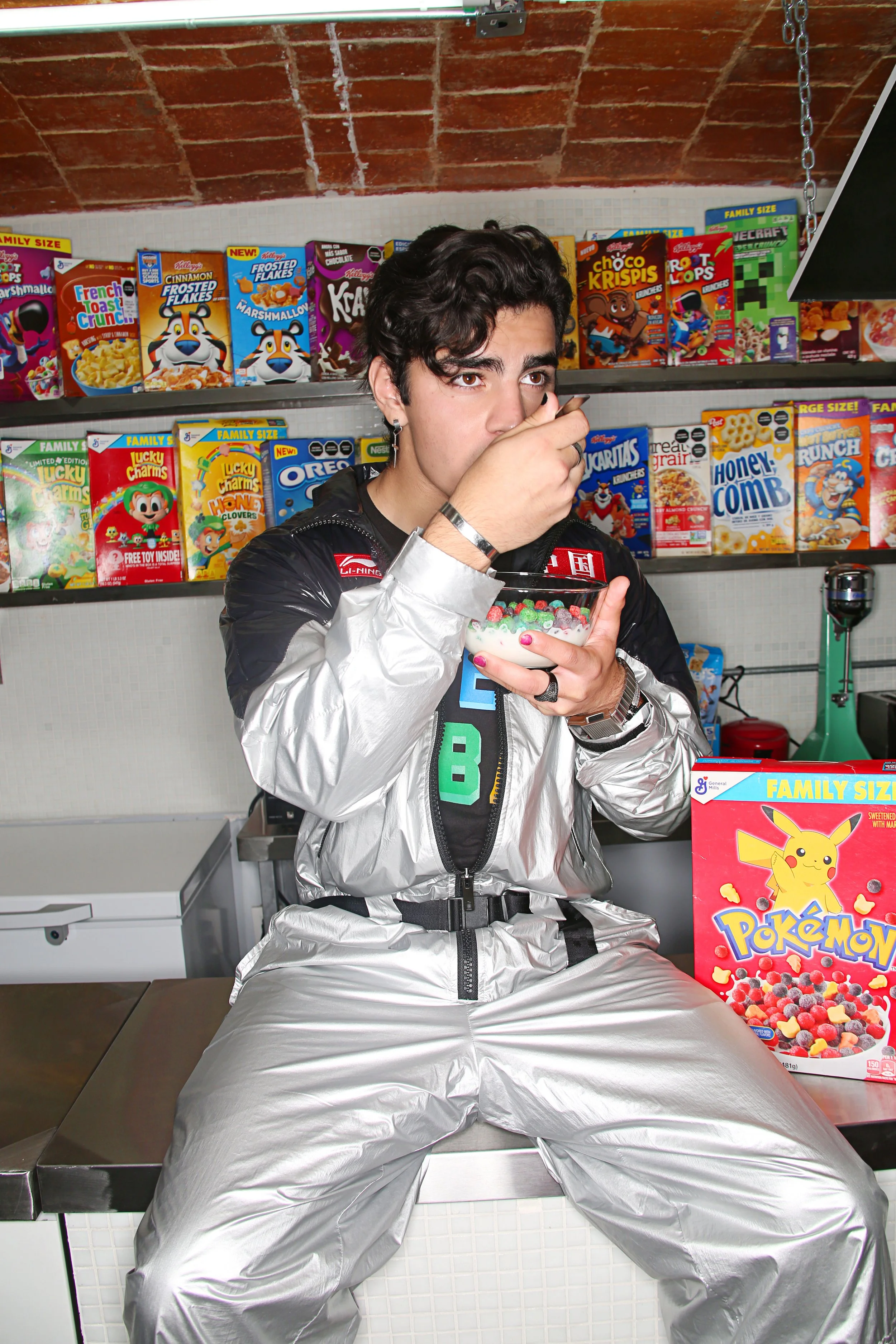 A person sitting on a scale, wearing a silver space suit, in front of a shelf of cereal boxes, holding a bowl of cereal and spoon, with a Pikachu cereal box on the counter.