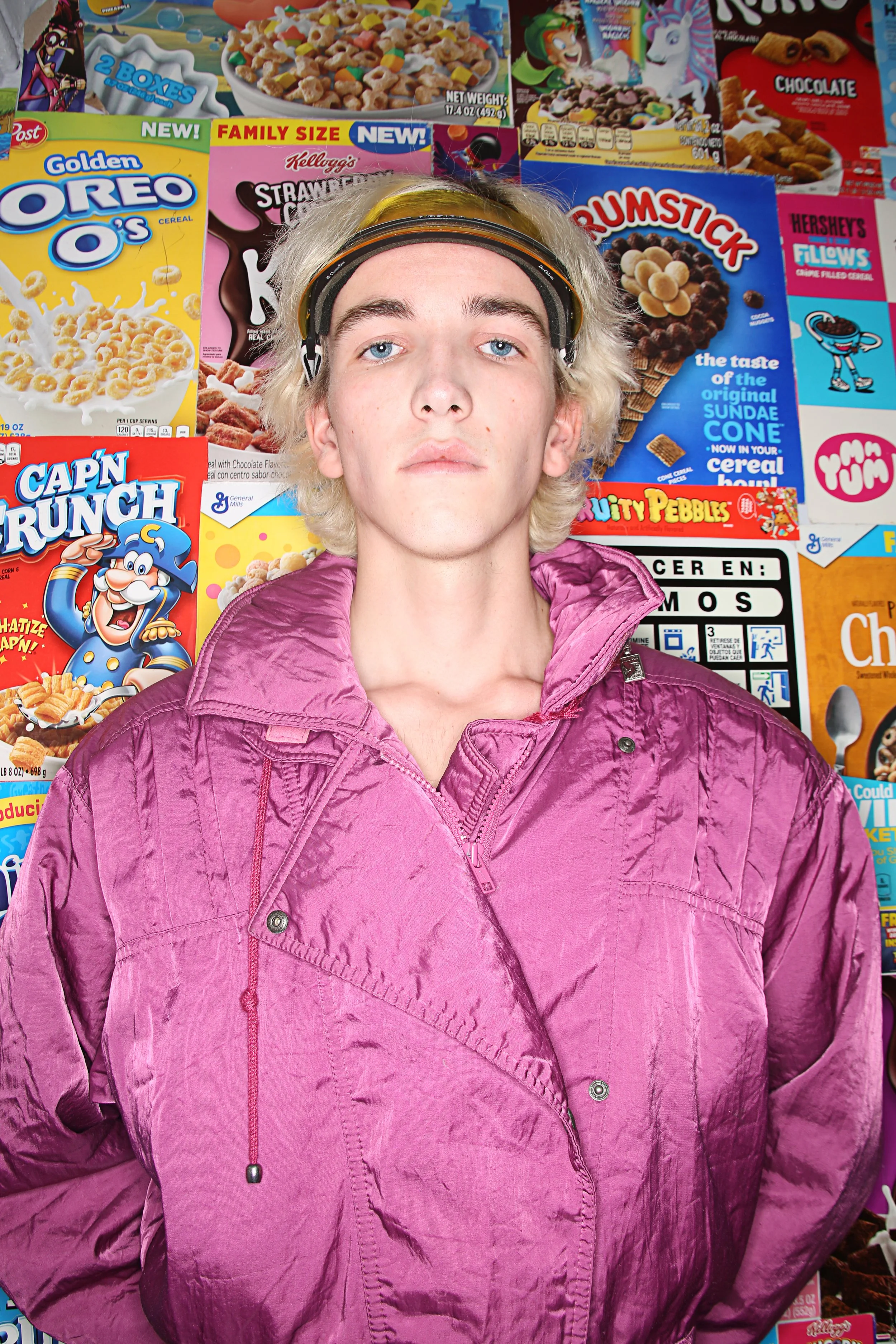 A young person with blond hair and blue eyes wearing a pink jacket, standing in front of a wall covered with cereal boxes and snack packaging.