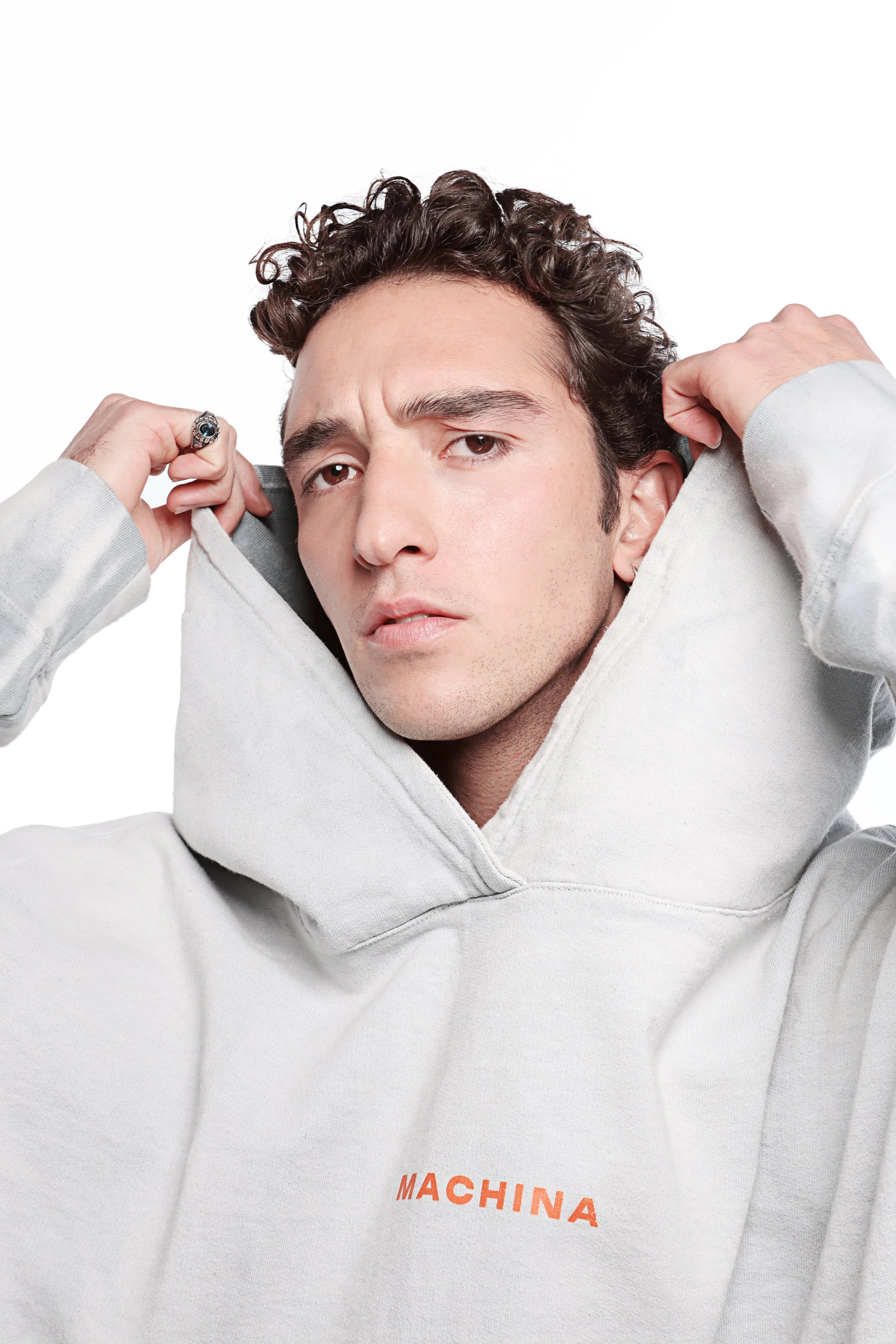 A man with curly hair and a ring on his finger is raising his arms behind his head and looking at the camera. He is wearing a light gray hoodie with the word "MACHINA" written in orange on the front.