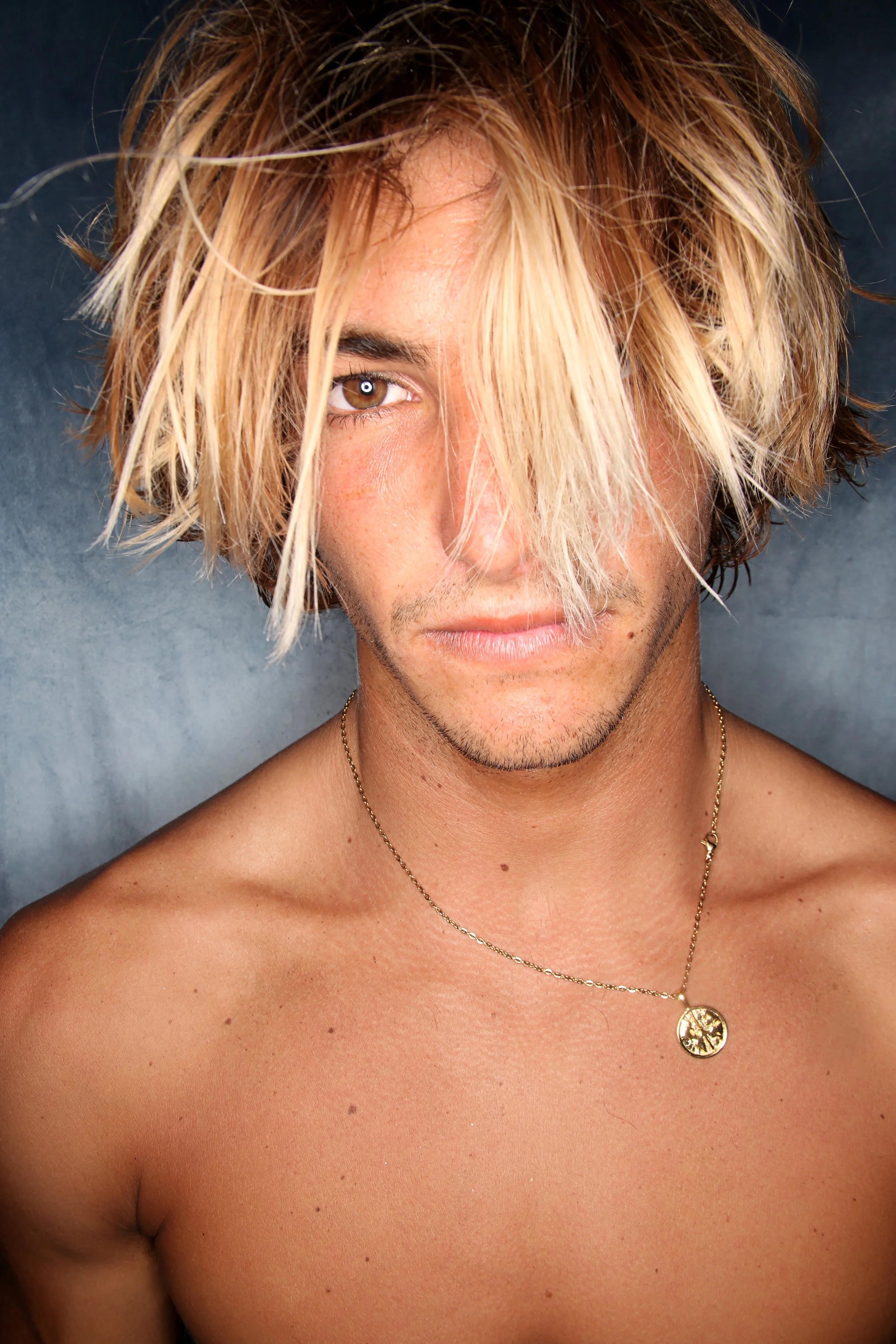Close-up of a shirtless man with tousled blonde hair, one eye visible, wearing a gold pendant necklace, against a dark background.