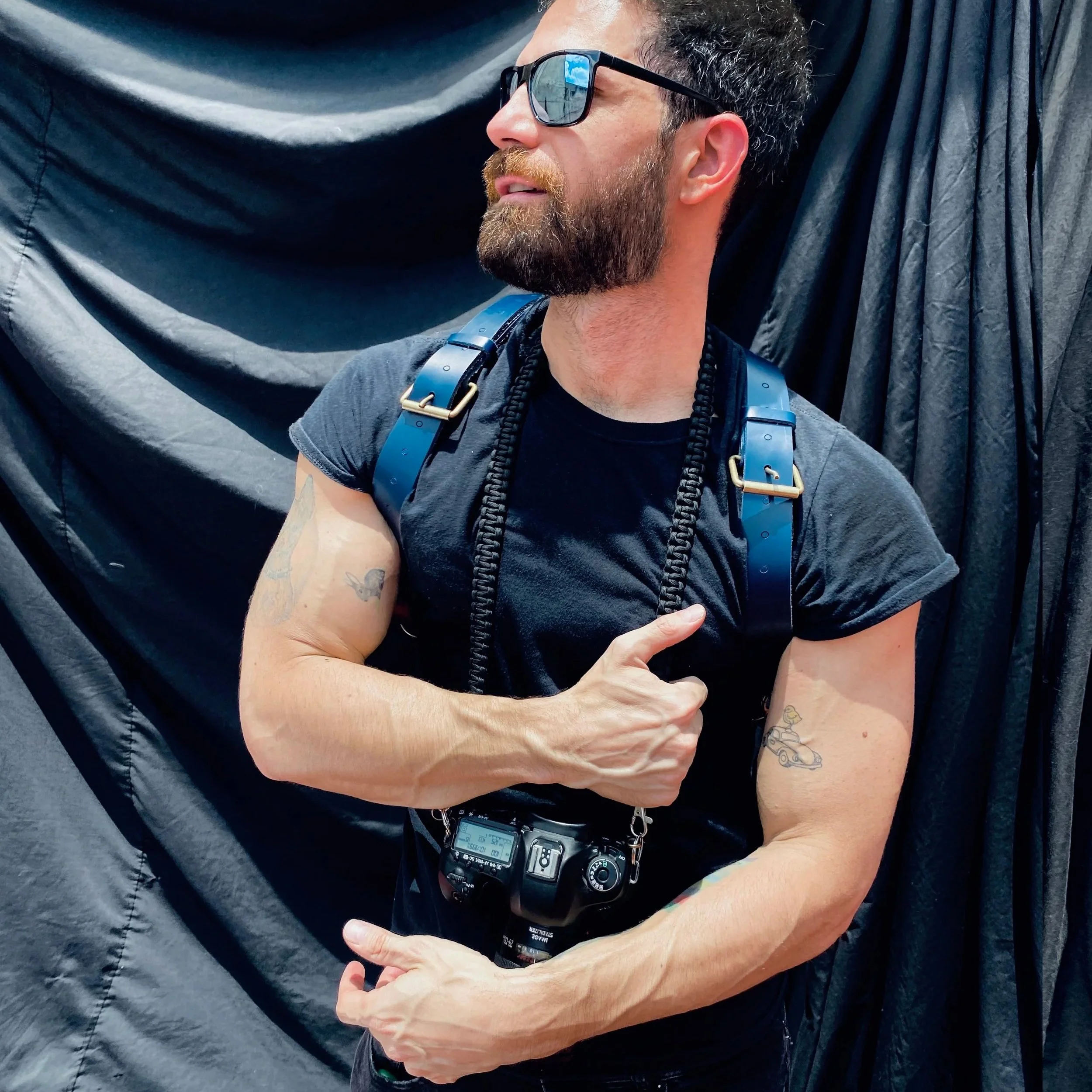 A man with dark sunglasses and a beard, wearing a black shirt and a camera strap around his neck, is hugging a person dressed in black with a dark quilted fabric background.