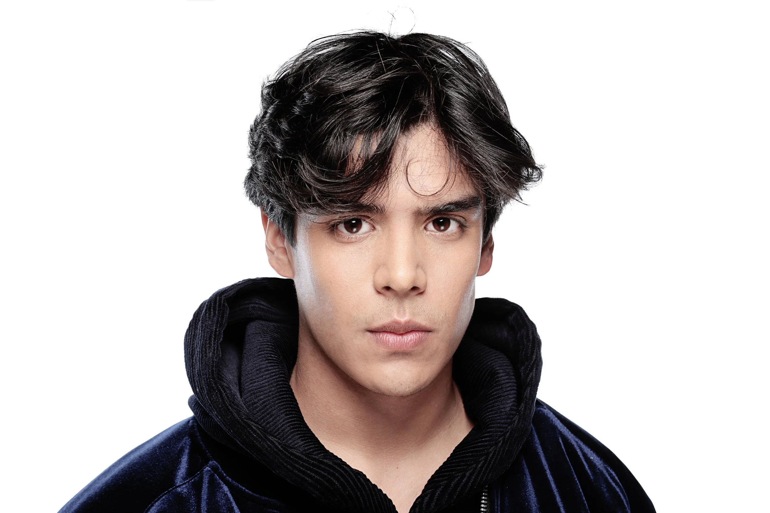 Close-up of a young man with dark, wavy hair, wearing a black velvet hoodie against a plain white background.