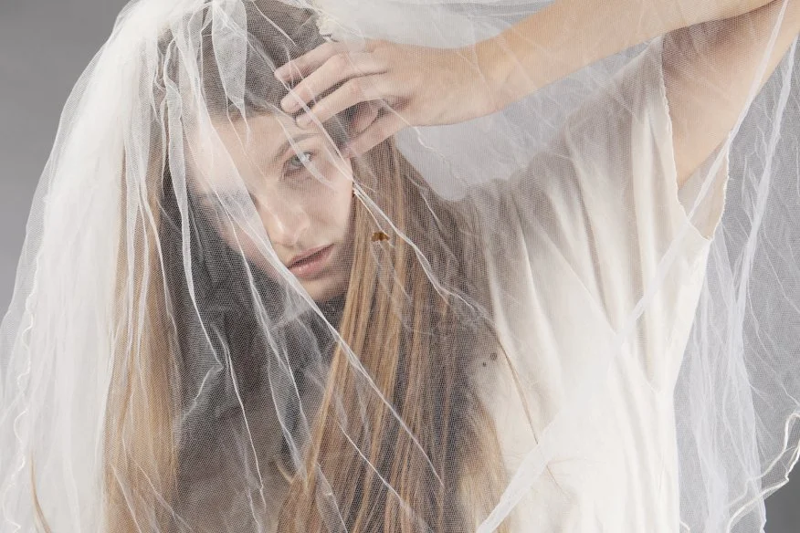 A woman with long light brown hair partially covered by a sheer white veil, looking directly at the camera with one hand touching her head, against a neutral background.