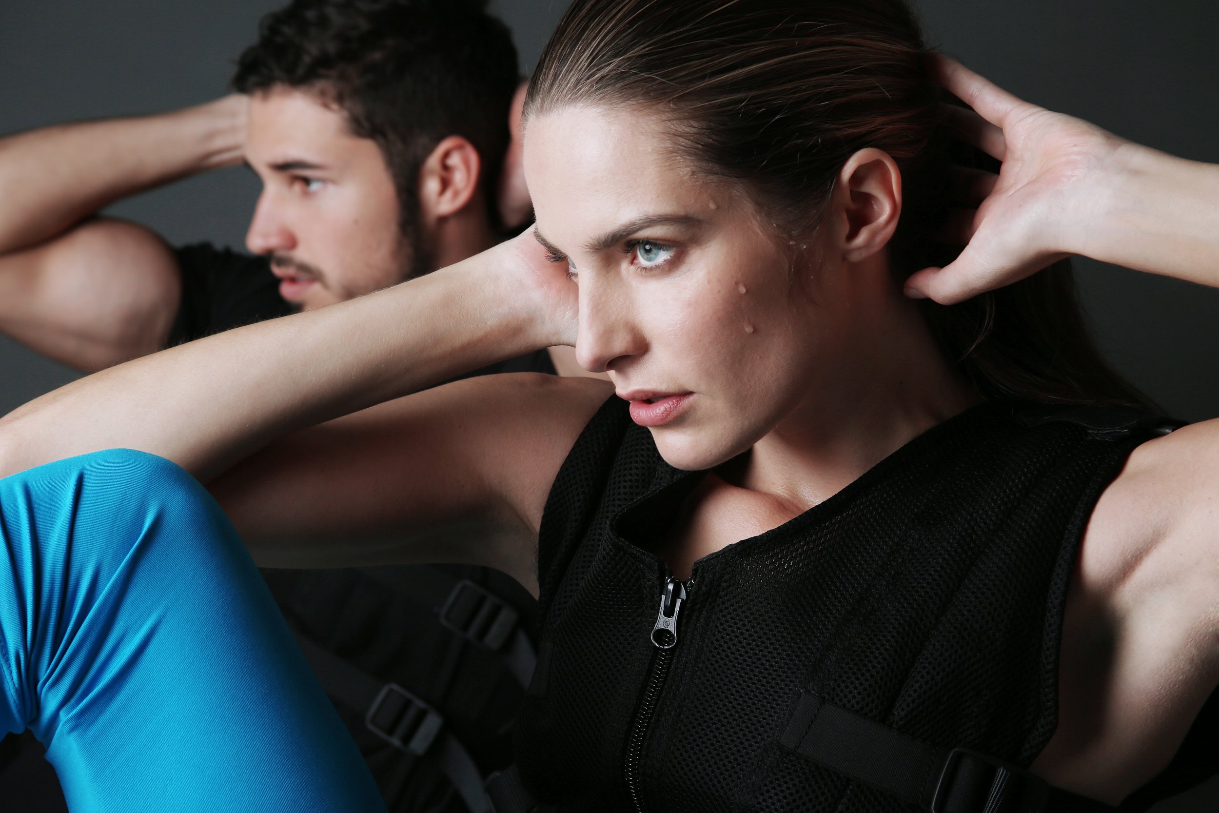A woman with wet hair and tears on her face, with a man behind her, both stretching their arms over their heads against a dark background.