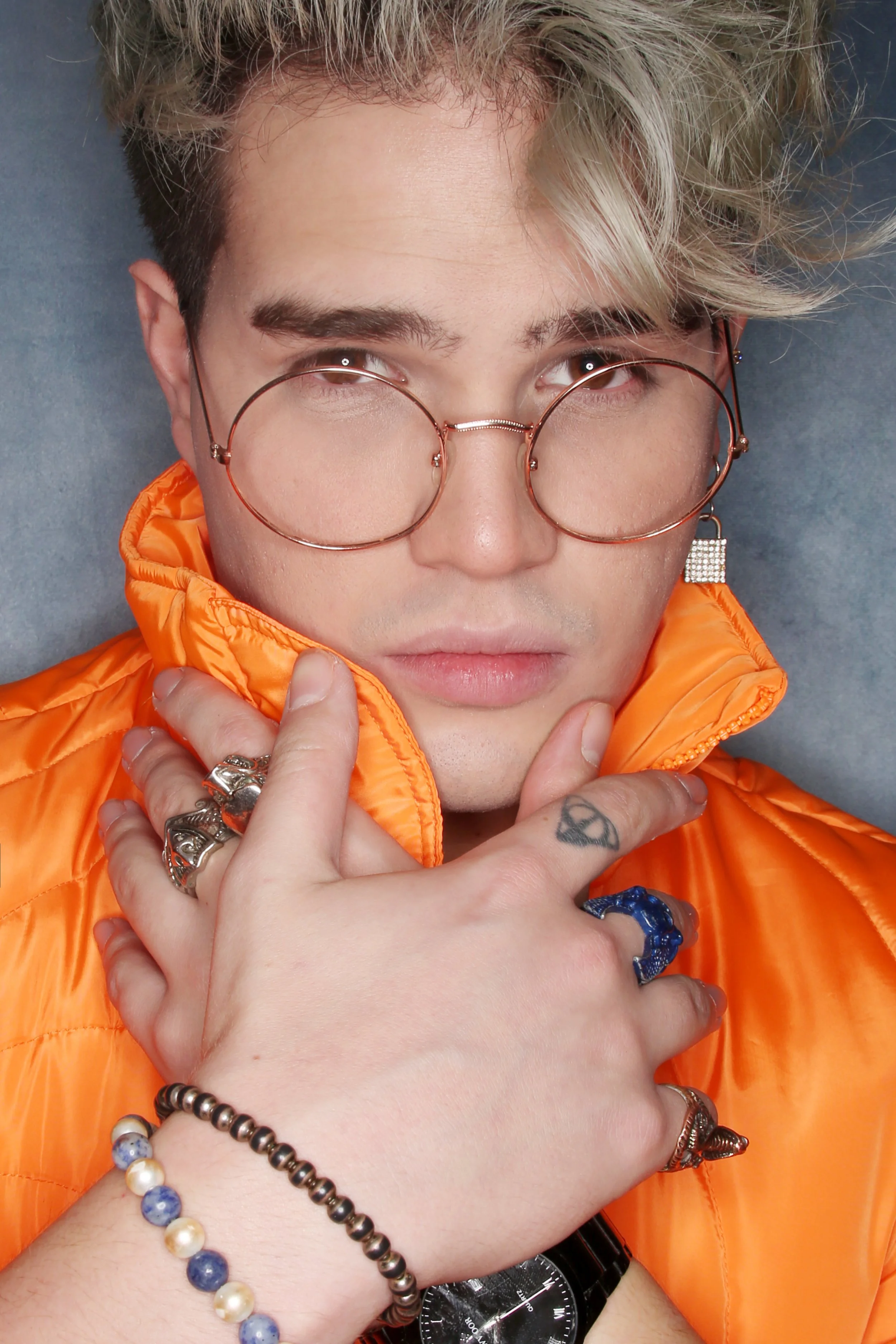 A person with blonde, wavy hair wearing round glasses, an orange puffy jacket, and various jewelry including rings, bracelets, and a watch, touching their chin with one hand, against a grey background.