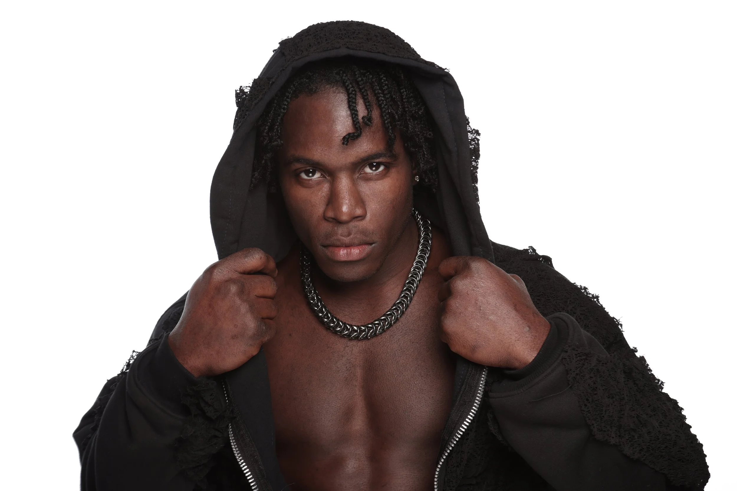 A young man with braided hair wearing a black hoodie, showing a necklace and holding the hoodie with both hands against a white background.