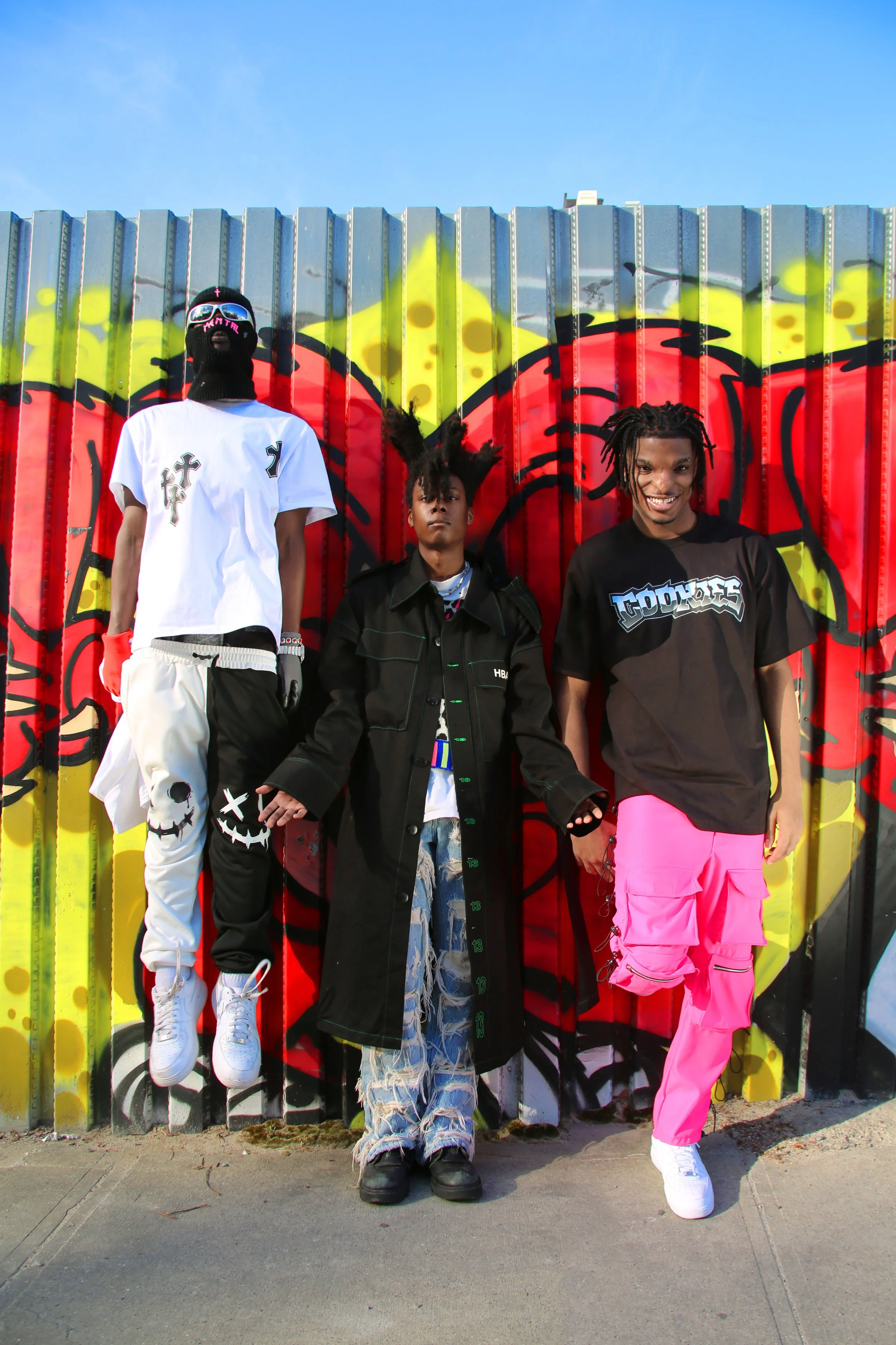 Three young men posing against a colorful graffiti wall outdoors. The man on the left is wearing a white oversized T-shirt, white pants with graphic designs, a black face mask, sunglasses, and white sneakers. The man in the middle has dreadlocks, is 