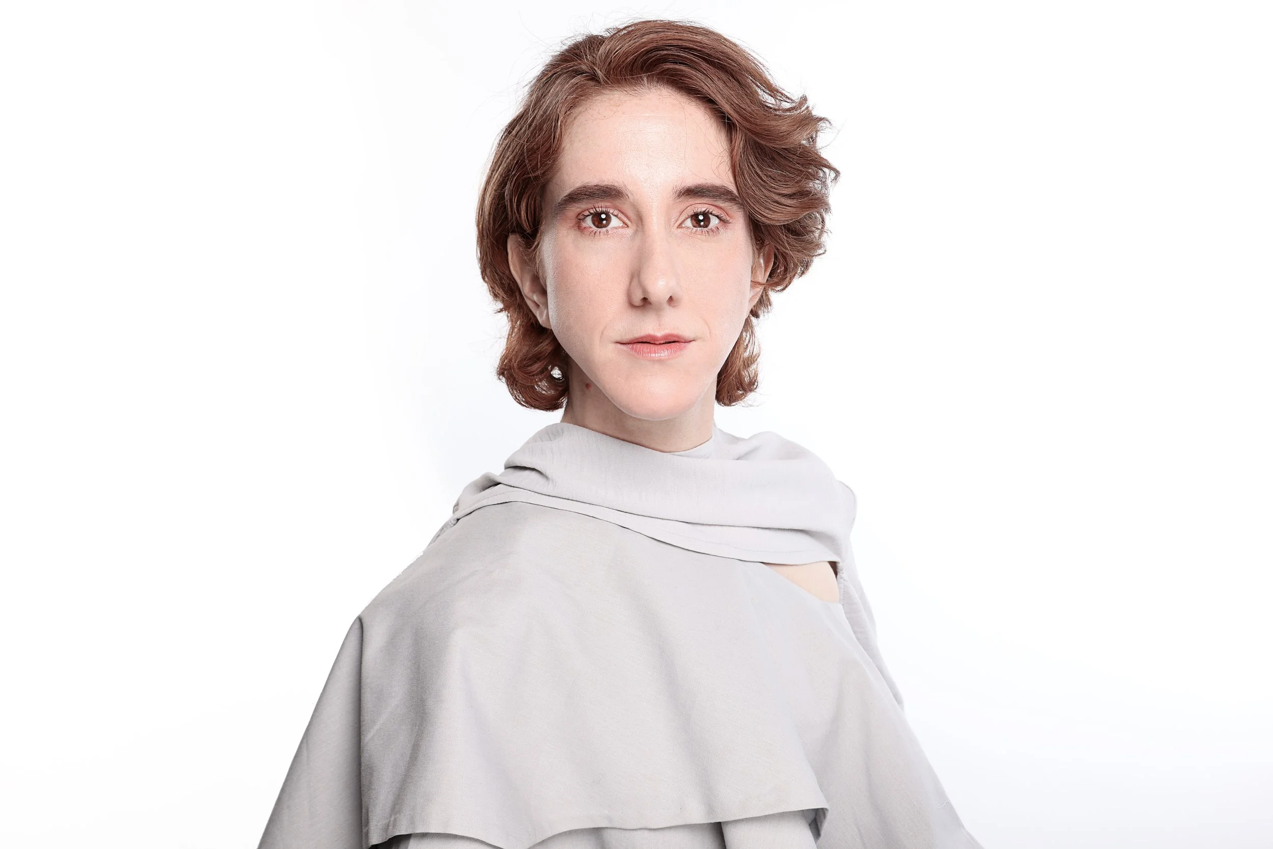 A portrait of a woman with short, wavy auburn hair wearing a light gray top with a draped neckline against a white background.
