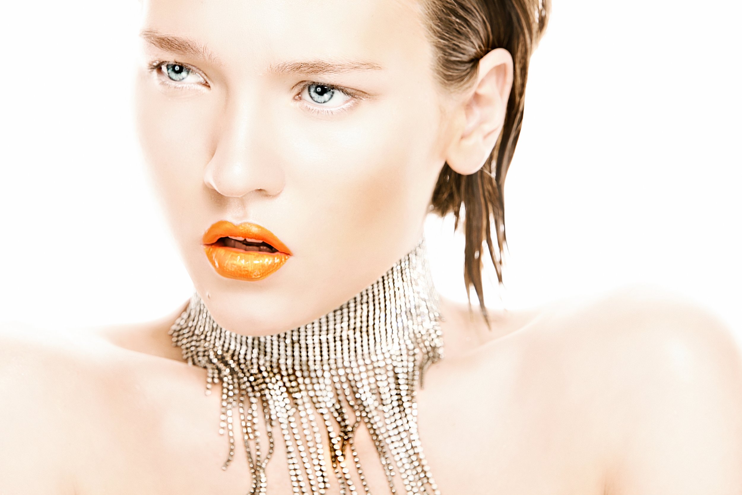 Close-up of a woman with short brown hair, blue eyes, orange lipstick, and wearing a shiny silver chainmail necklace.