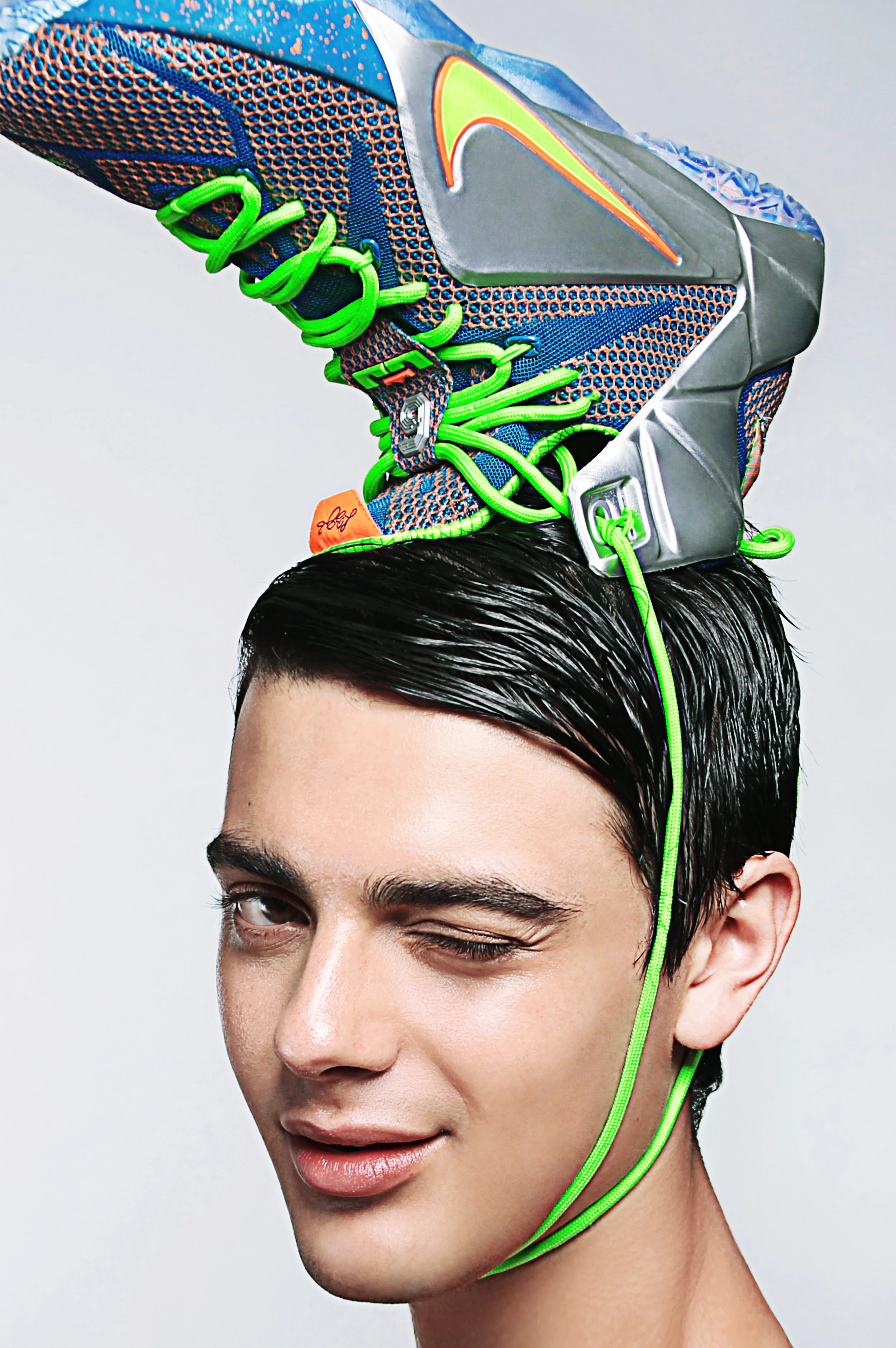 A young man with black hair, partly slicked back, wears a futuristic sneaker on his head, with neon green laces and a gray Nike swoosh logo, against a plain white background.