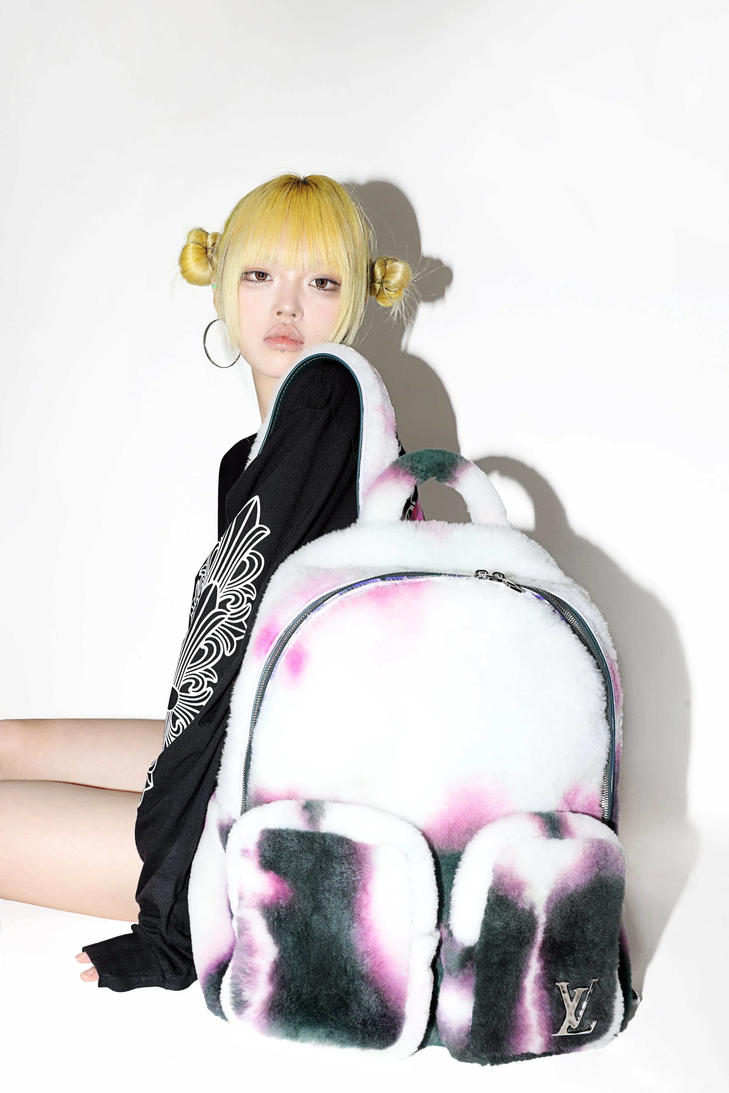 Young woman with blonde hair styled in buns, wearing large hoop earrings, holding a colorful plush Louis Vuitton bag.