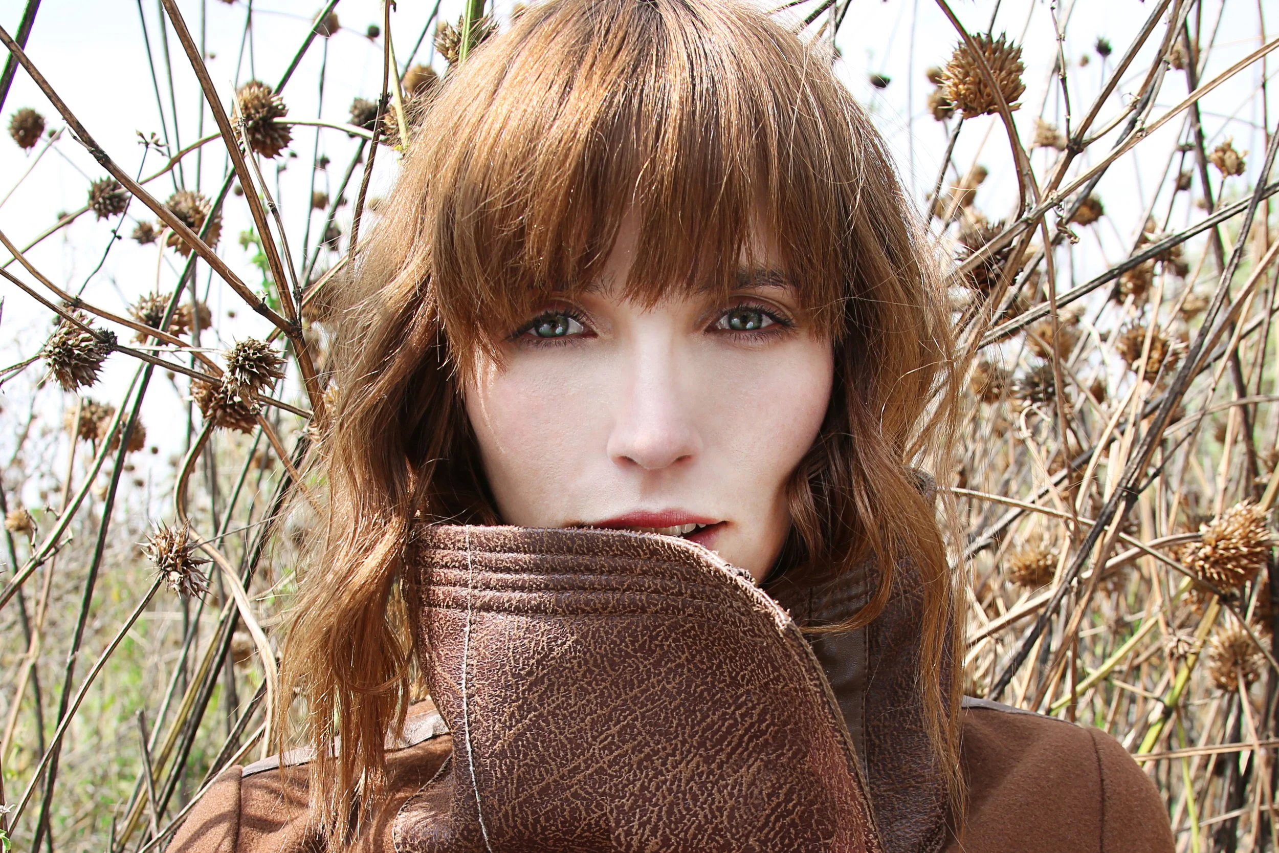 A young woman with red hair and green eyes looking into the camera while standing outdoors among dry, thorny plants.