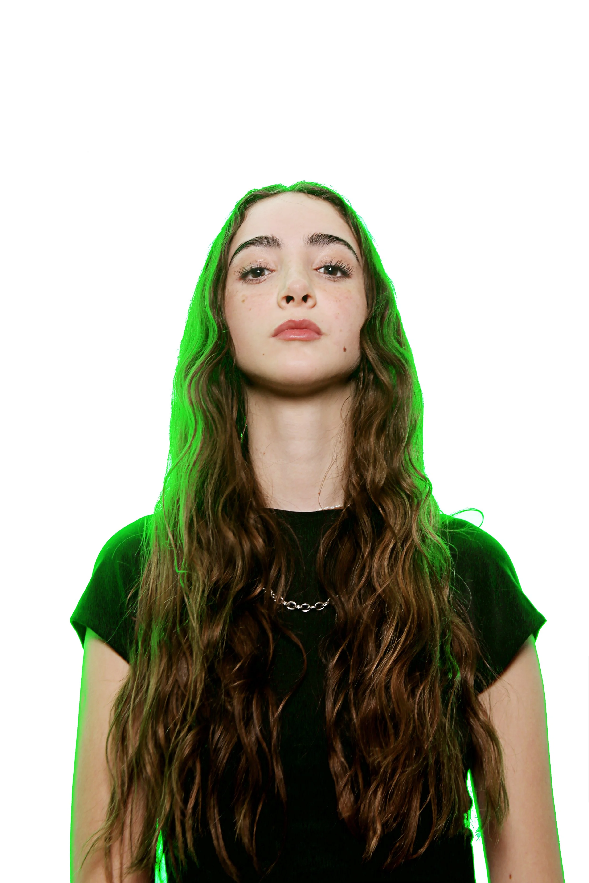 A young woman with long wavy brown hair, wearing a black shirt and chain necklace, is looking directly at the camera with a serious expression against a plain white background.