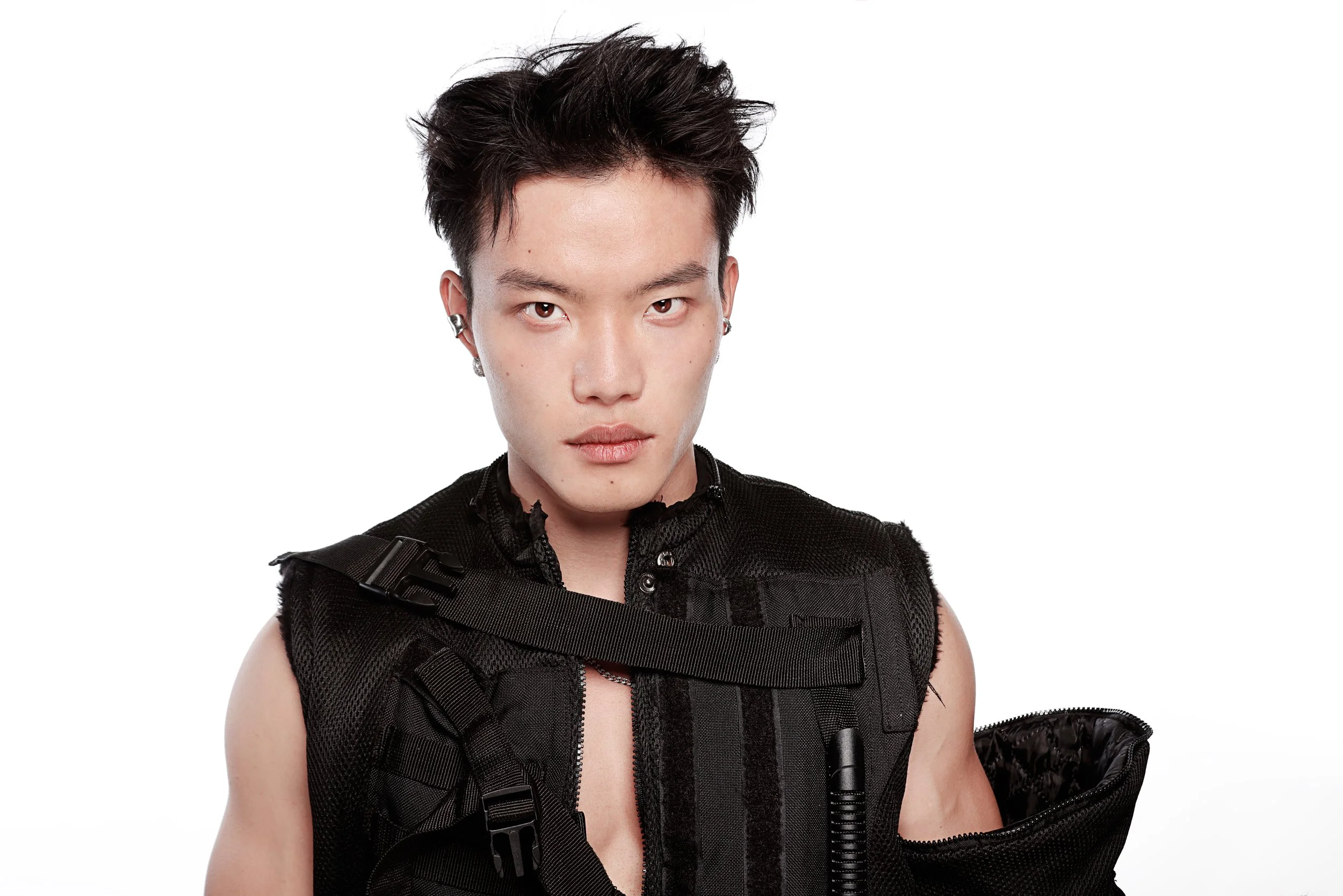 A person with short dark hair wearing a black vest with straps and holding a jacket, standing against a plain white background.