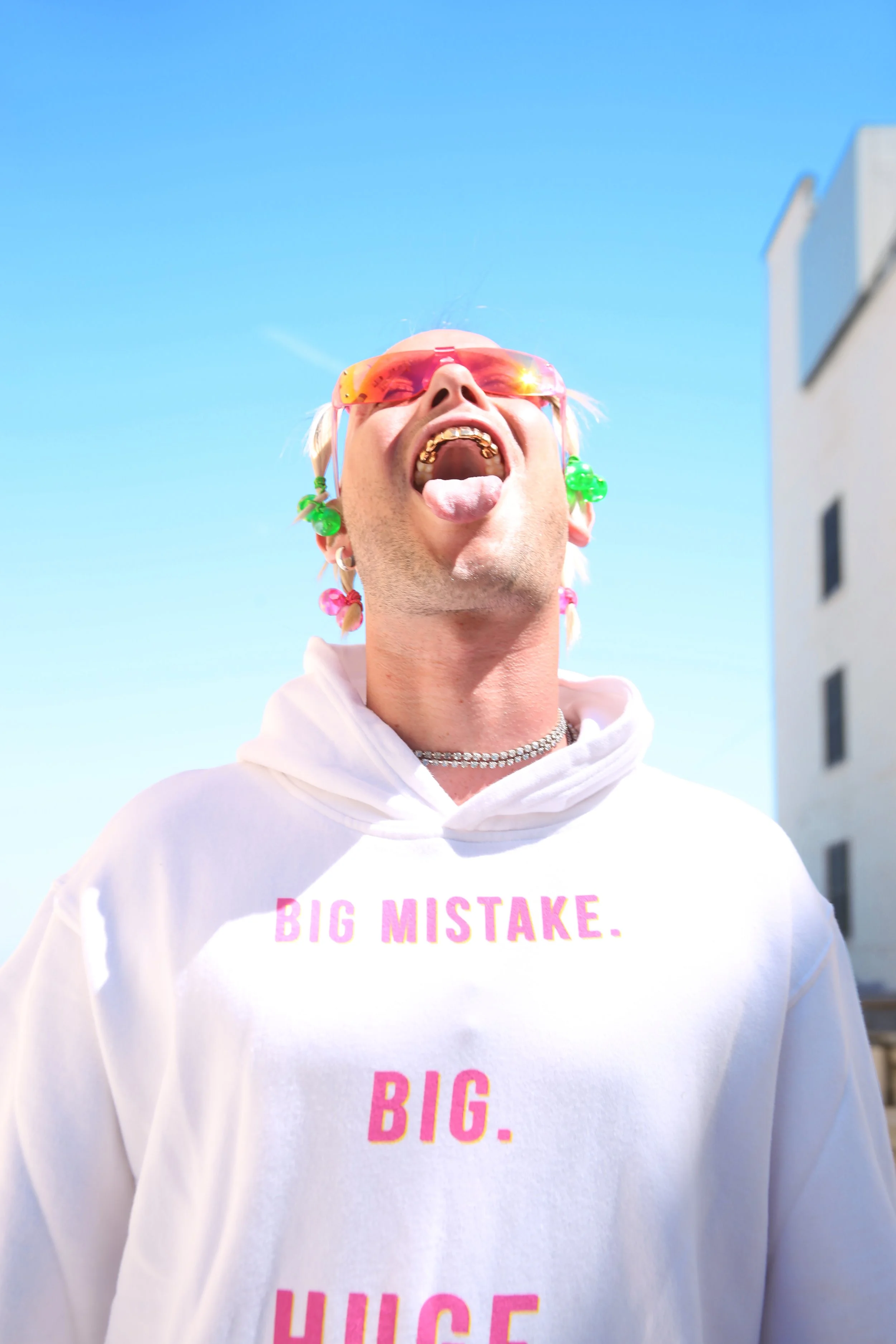 Close-up of a person with platinum blonde hair, wearing orange sunglasses, tongue sticking out, and a white hoodie with pink text reading 'BIG MISTAKE'.