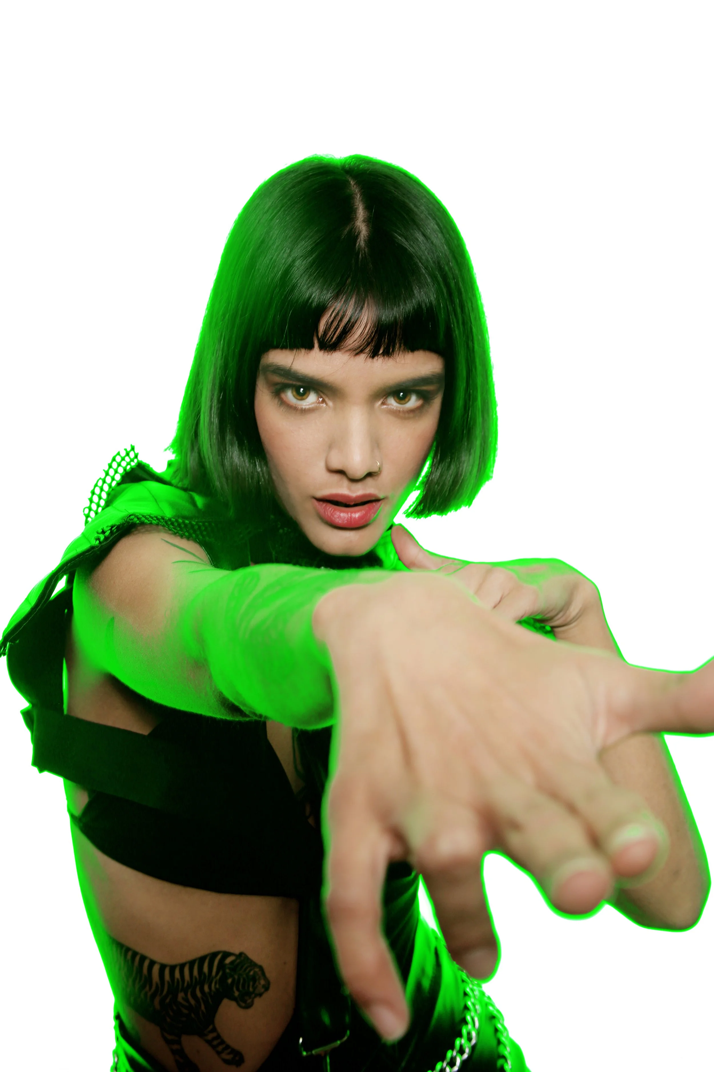 A woman with short black hair and a nose piercing points her finger toward the camera. She is wearing a black top with a tiger tattoo visible on her arm and has green lighting highlighting her hair and arm.